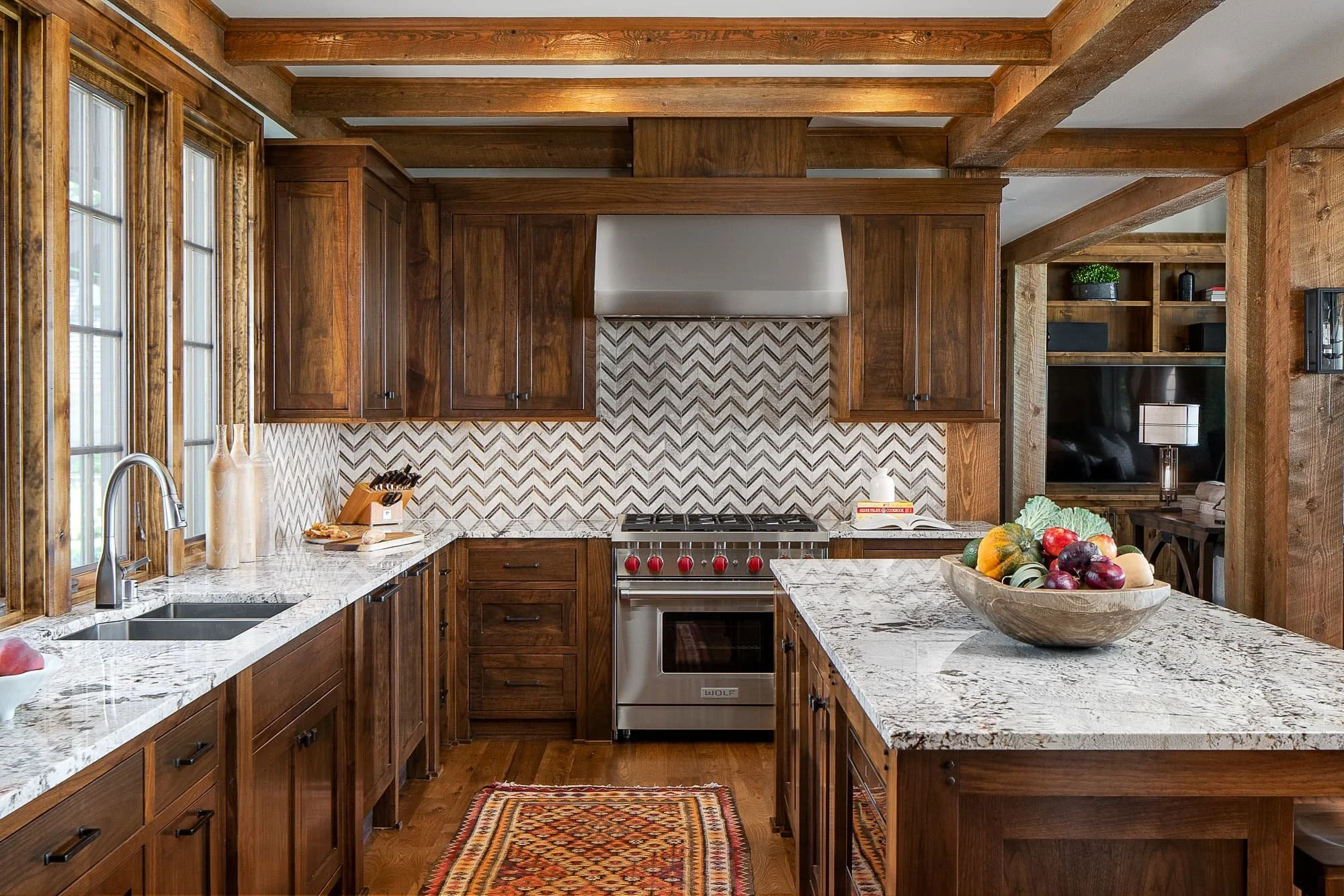 Warm kitchen with exposed wood beams, stained cabinetry, patterned tile backsplash, stone countertops, and a large island with a fruit bowl.