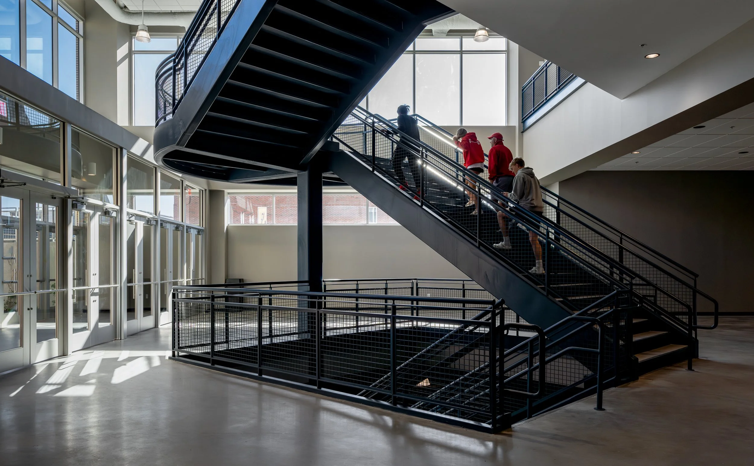 Bright atrium with black steel staircase, glass walls, concrete floors, and people on the stairs.