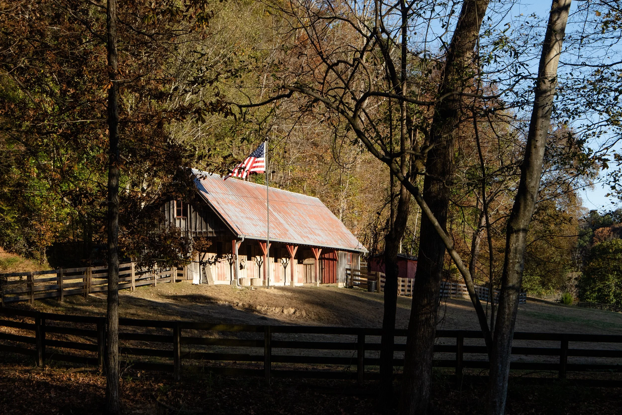 Weathered barn with a metal roof and American flag, set behind wood fencing on a sloped rural property.