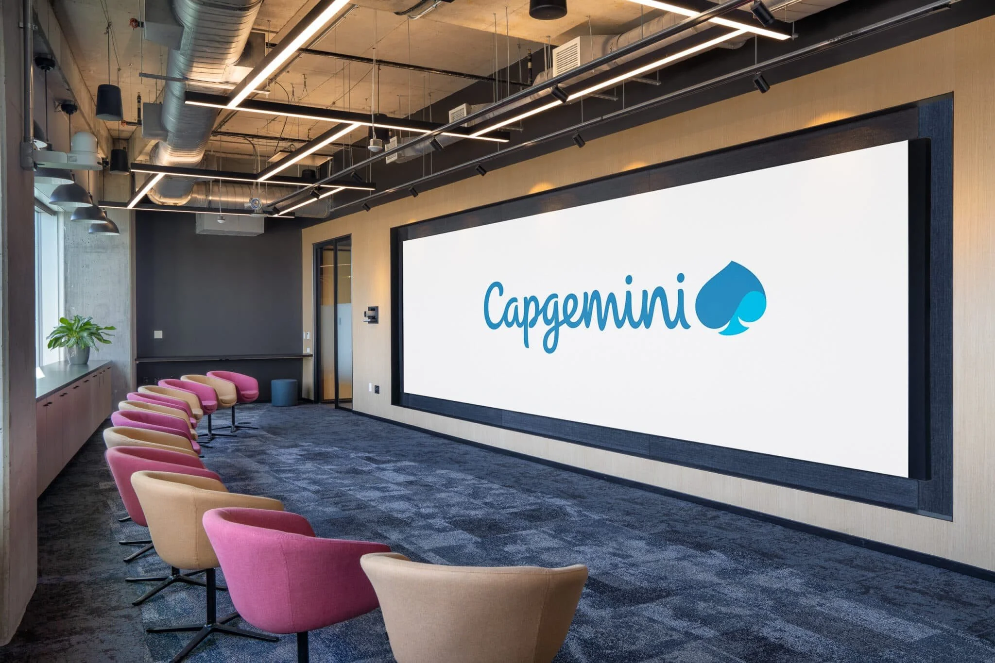 Presentation room with a large Capgemini display wall, rows of colorful lounge chairs, and dark carpet.
