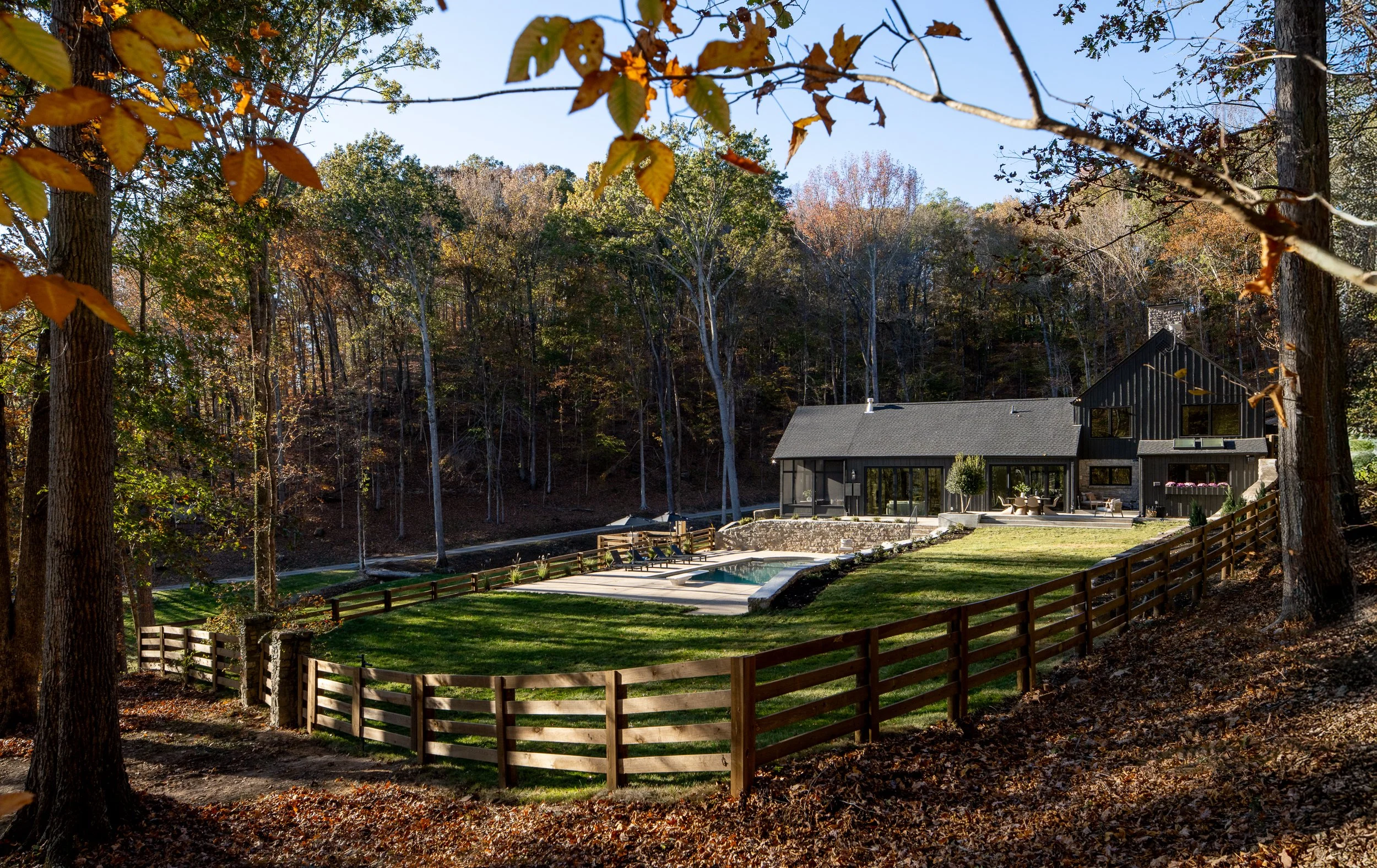 A modern house with large windows and a dark exterior, surrounded by a well-maintained yard with a swimming pool and patio furniture, set against a backdrop of trees with autumn leaves.