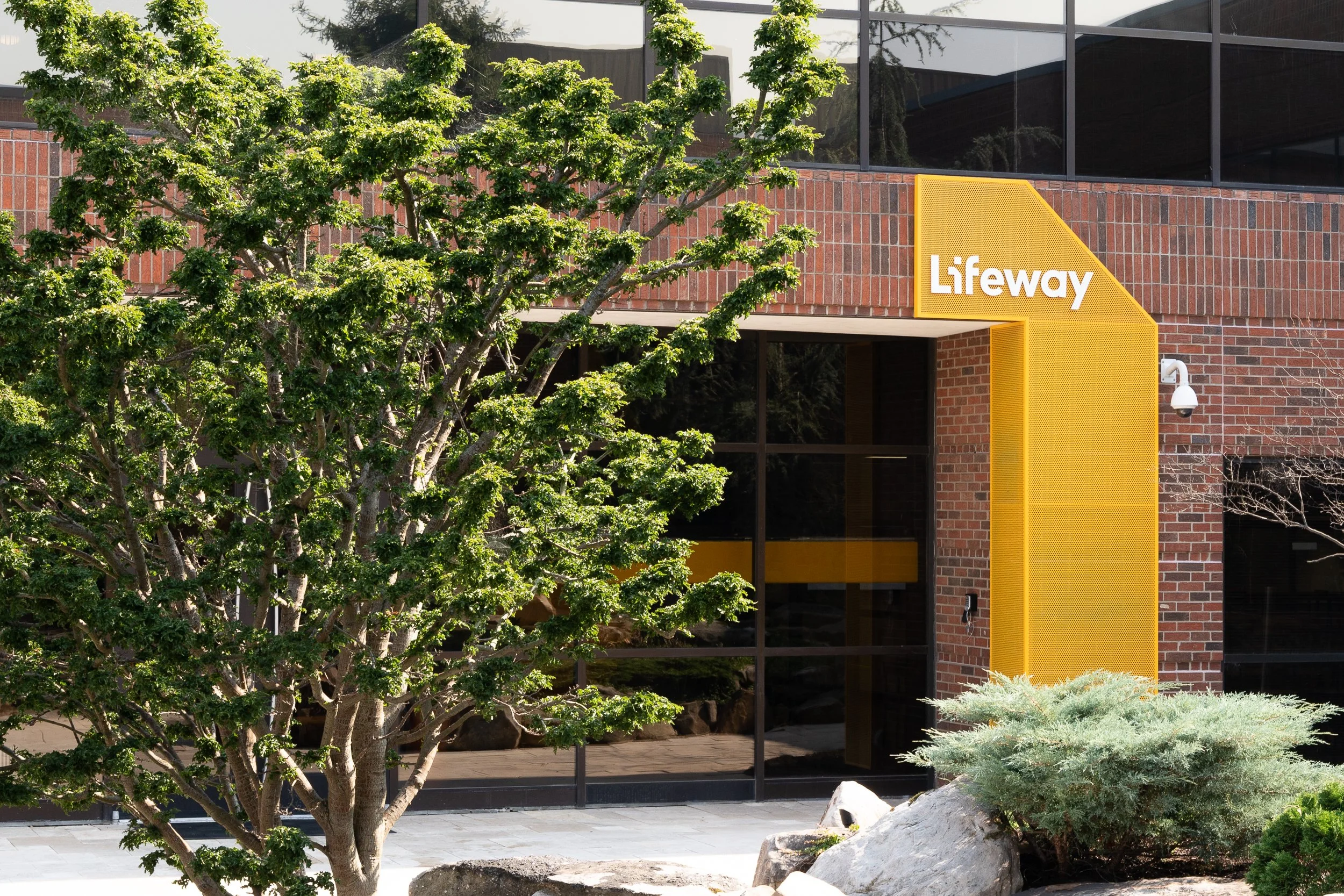Lifeway building entrance with a yellow architectural canopy framed by brick, glass, and landscaping.