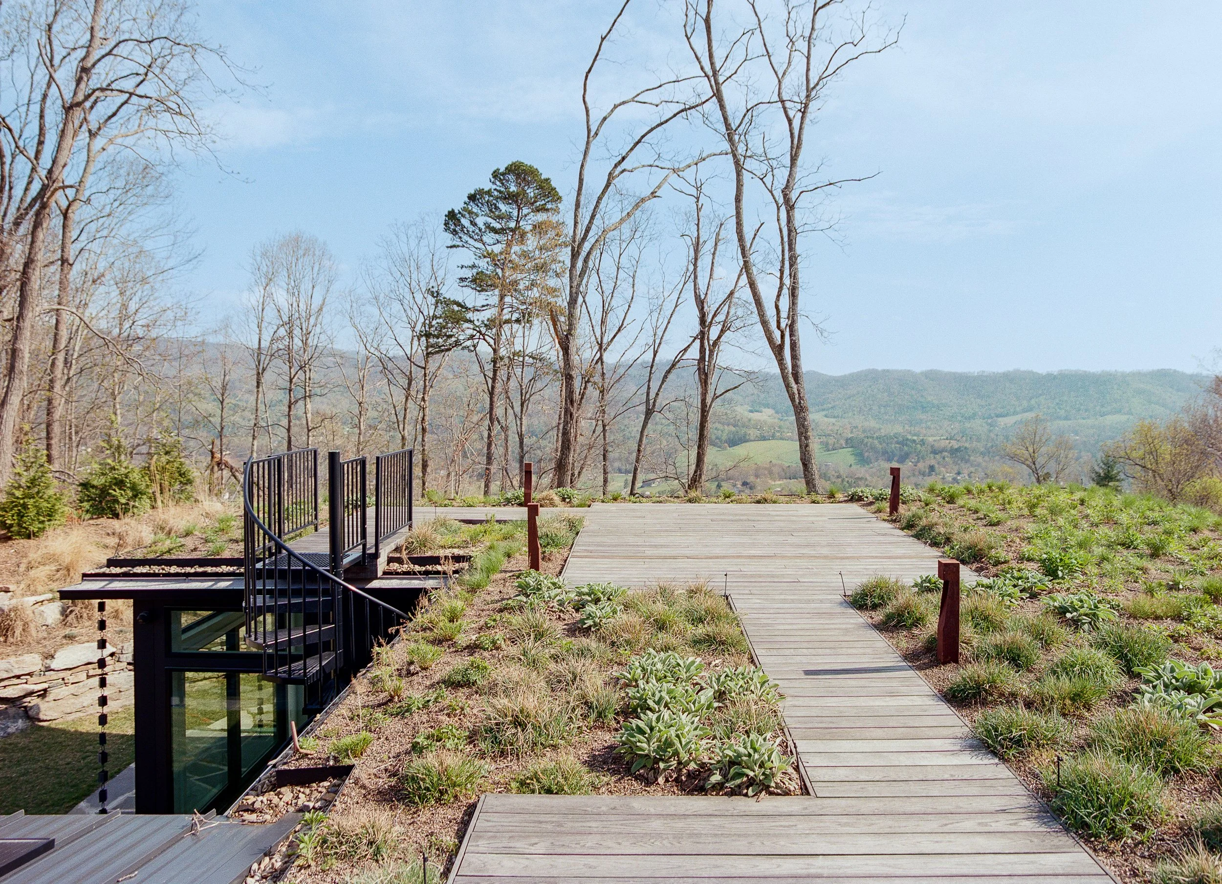 A wooden pathway in a natural park or garden with some sparse trees and mountain hills in the background, under a partly cloudy blue sky.