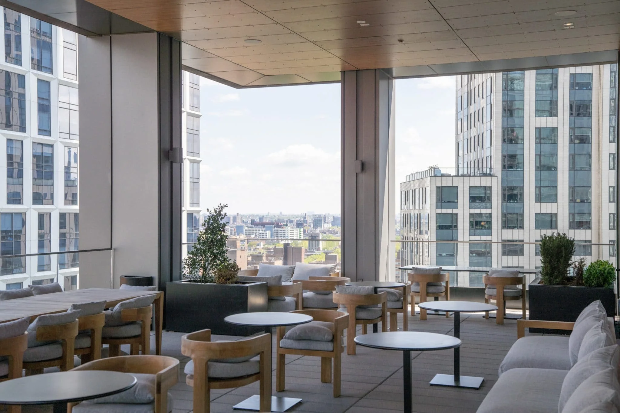 Covered outdoor lounge with wood chairs, round tables, planters, and views between surrounding high-rise buildings.