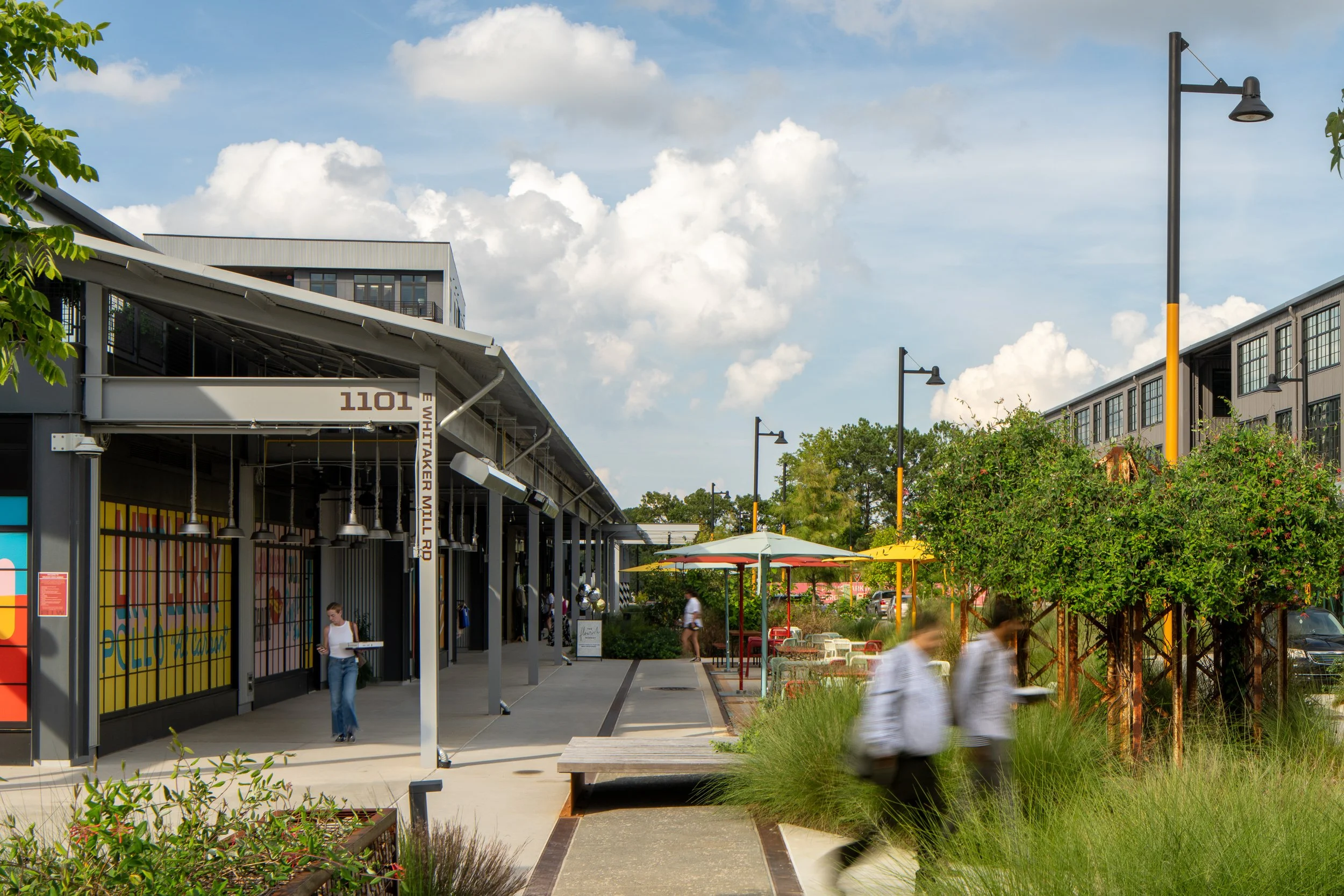 An outdoor shopping or dining area with colorful umbrellas, benches, and a few people walking. Modern buildings and street lamps are visible under a partly cloudy sky.