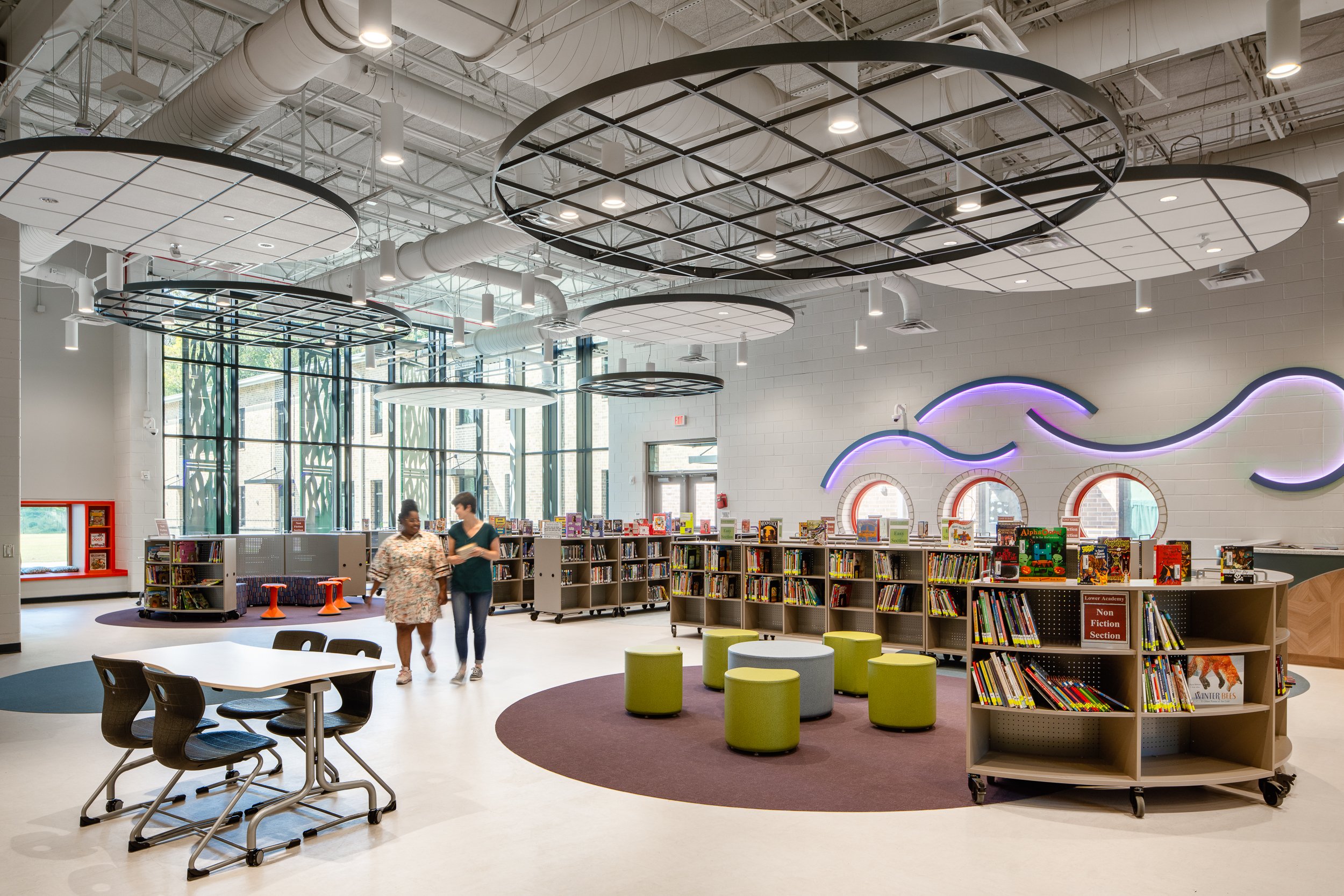Bright school library with circular suspended ceiling features, colorful seating, bookshelves, and two people walking through the space.