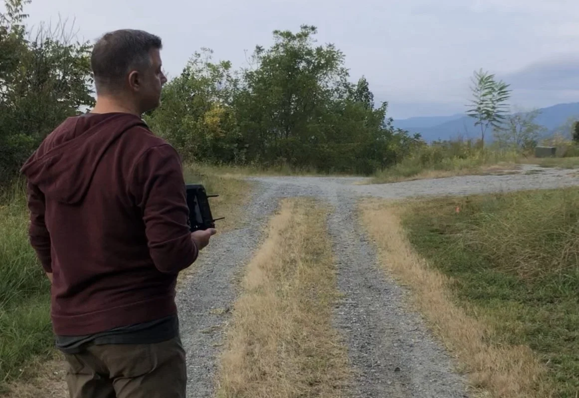 A man with a drone controller standing on a gravel dirt path in a natural area with trees and mountains in the background.
