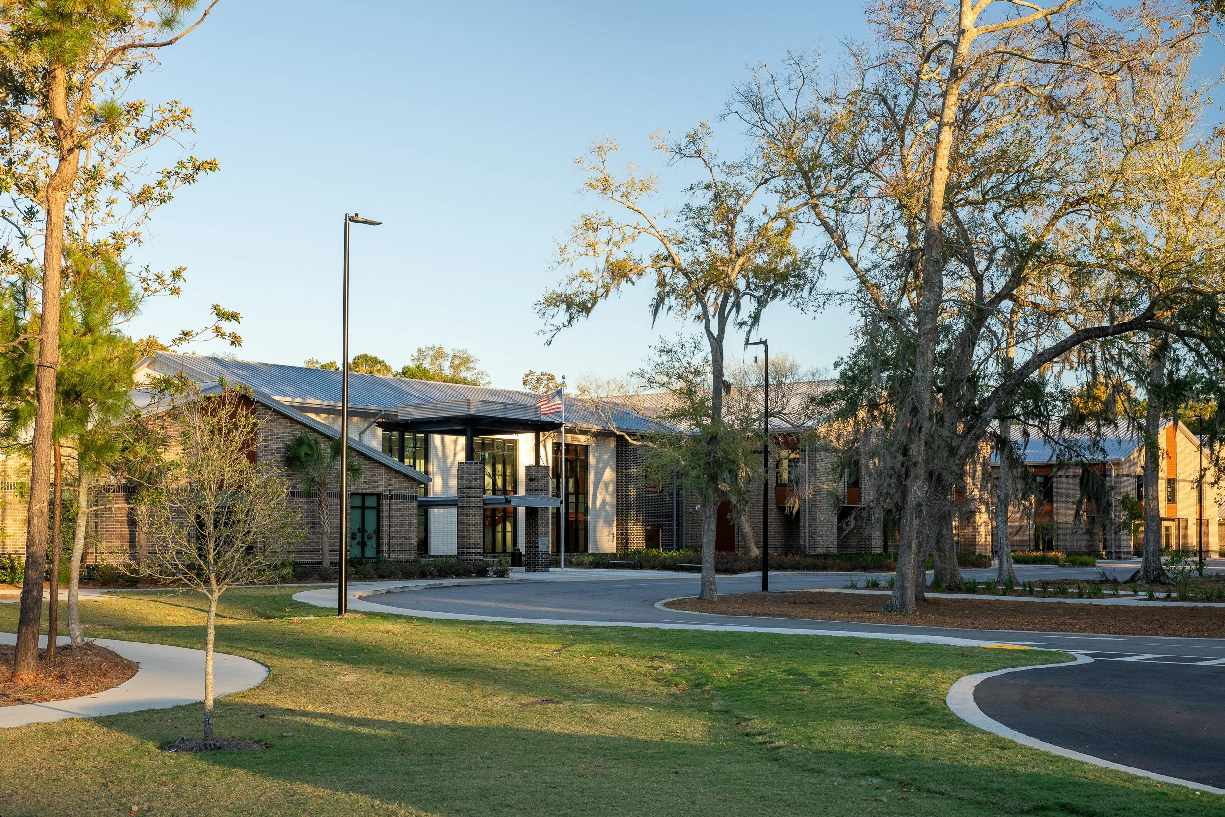 Wide view of the school campus with curving walkways, lawn, brick buildings, live oaks, and soft morning light across the site.