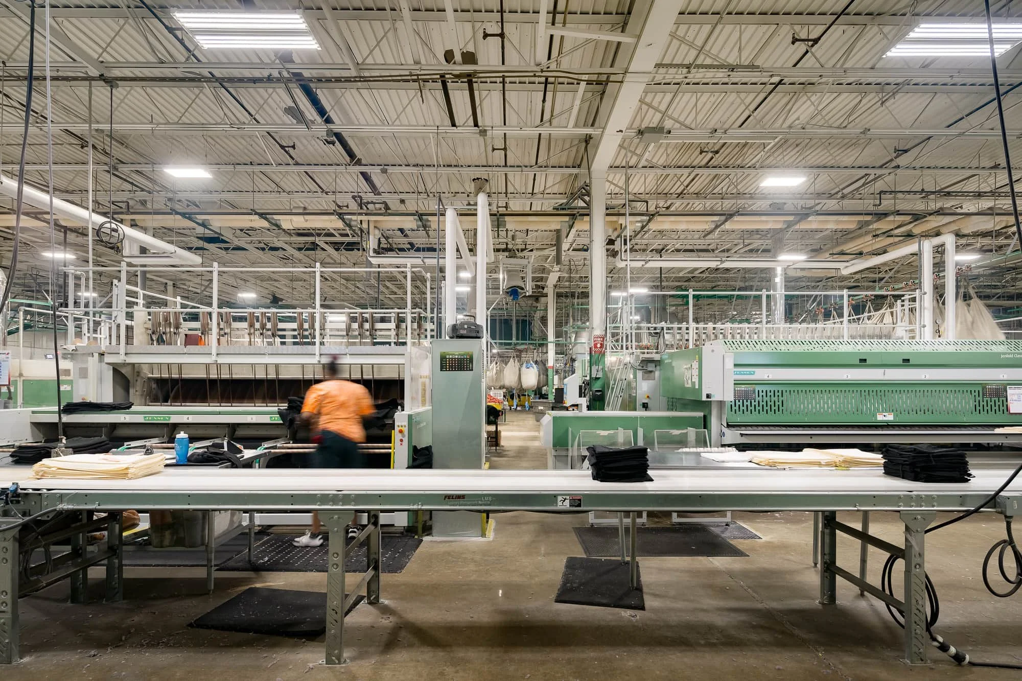 Large manufacturing facility with conveyor tables, industrial machinery, overhead utilities, and a worker moving through the production area.