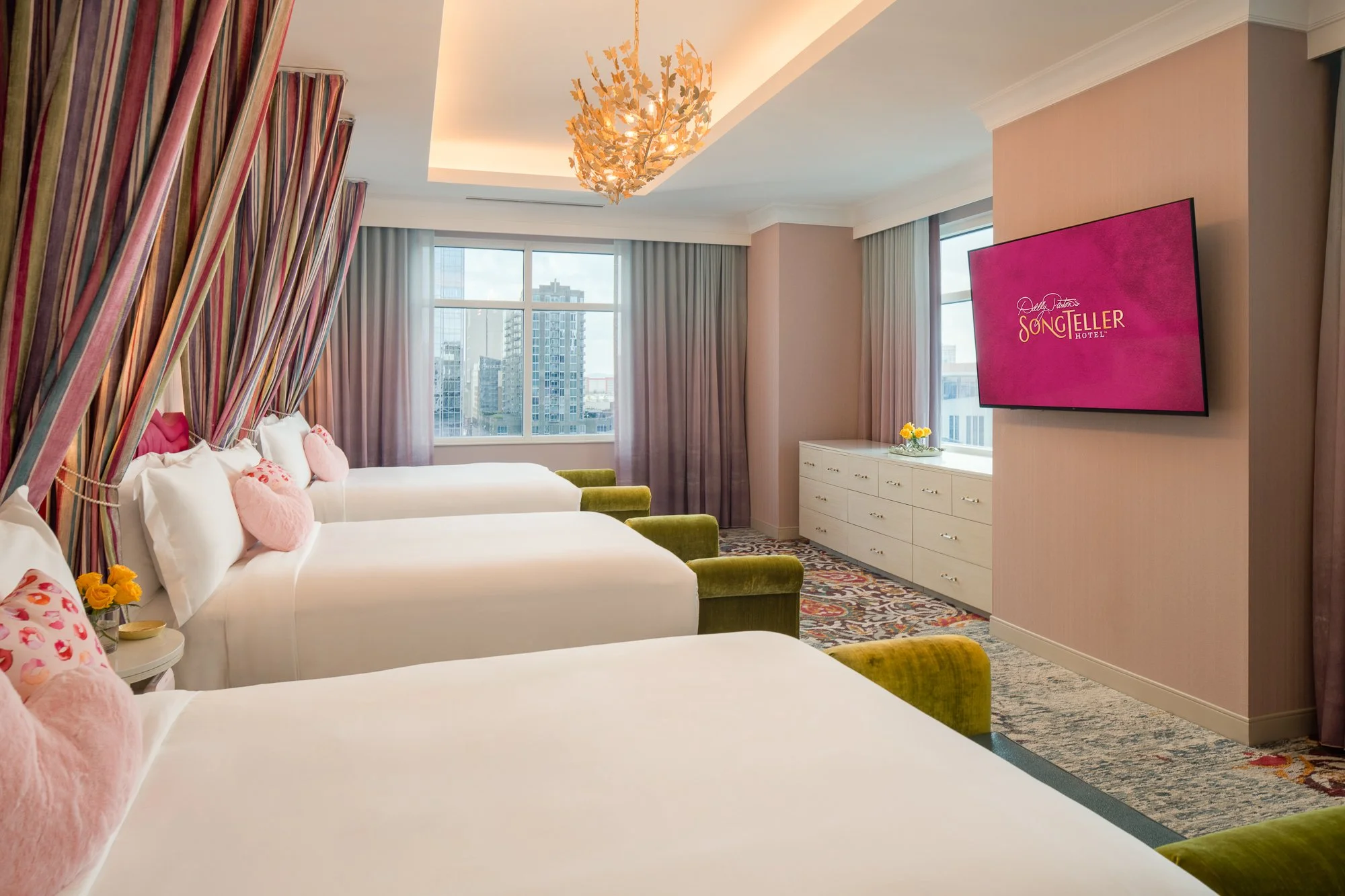 Three-bed hotel suite with white bedding, layered canopy drapery, green benches, chandelier, large windows, and wall-mounted television.