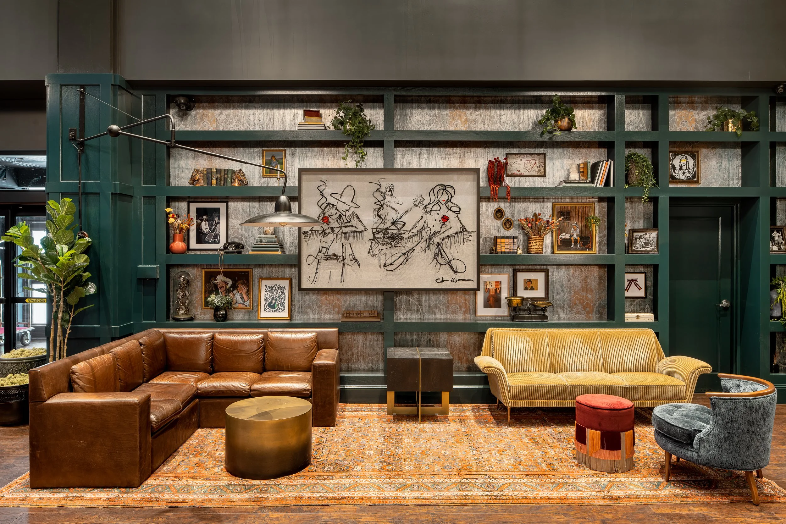 A cozy, well-decorated living room featuring brown leather sofa, yellow upholstered armchair, small round coffee tables, patterned area rug, and a wall with various framed photos, artwork, books, plants, and decorative items.