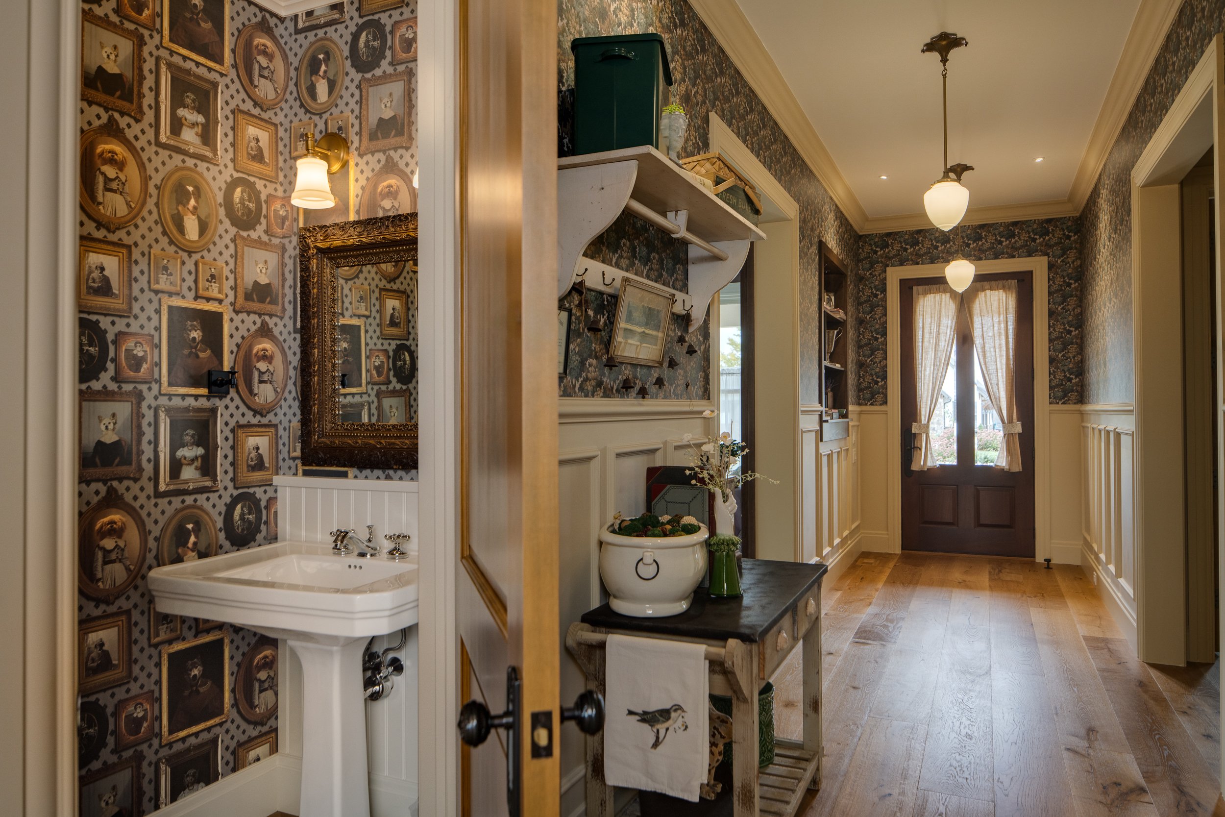 An interior view of a hallway with floral wallpaper, a door with curtains, and a vintage-style chandelier. The hallway includes a small table with decorative items and a wall-mounted shelf with picture frames.
