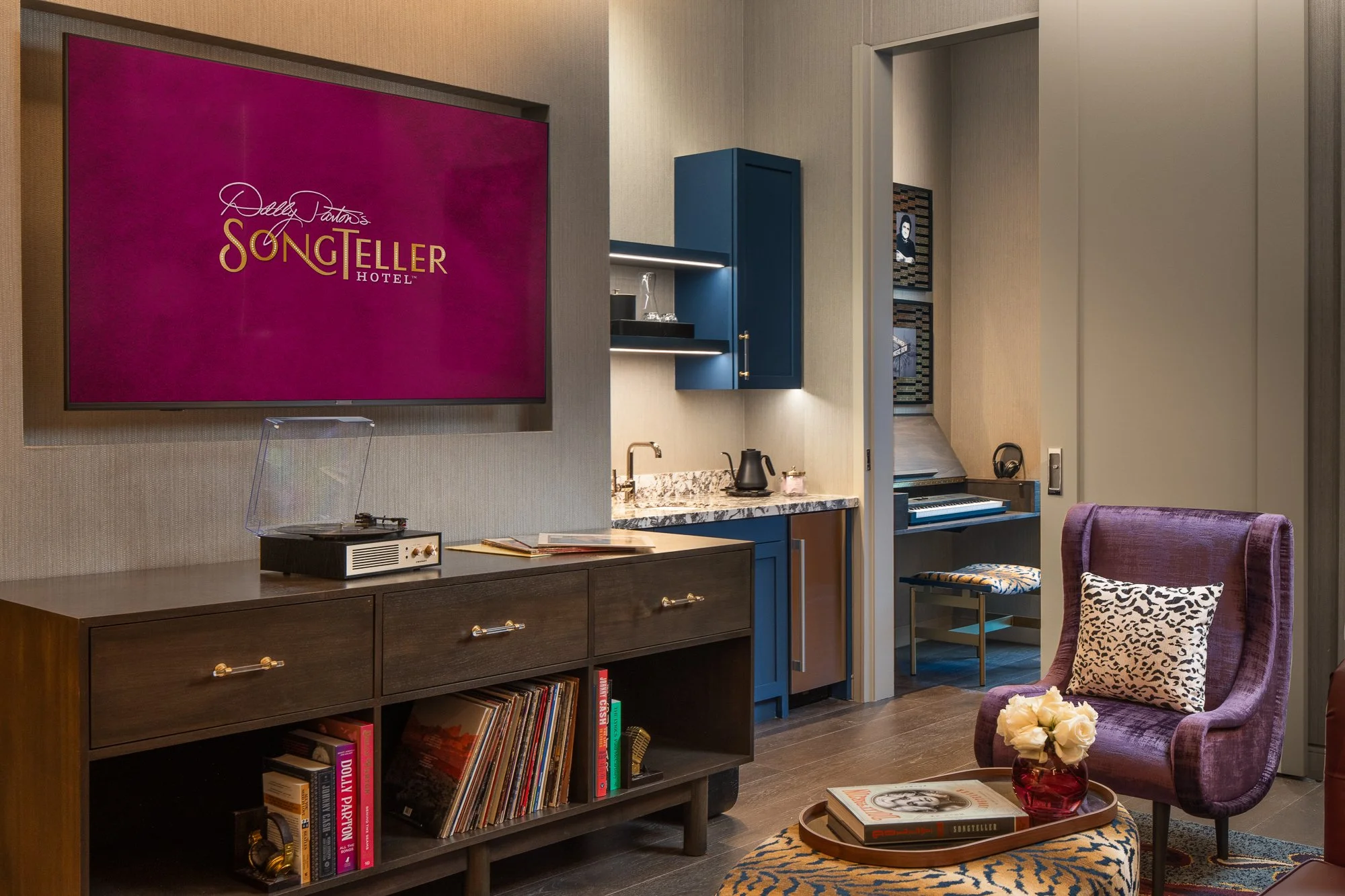 SongTeller Hotel suite living area with a record player, custom cabinetry, purple chair, television, and keyboard nook beyond the doorway.