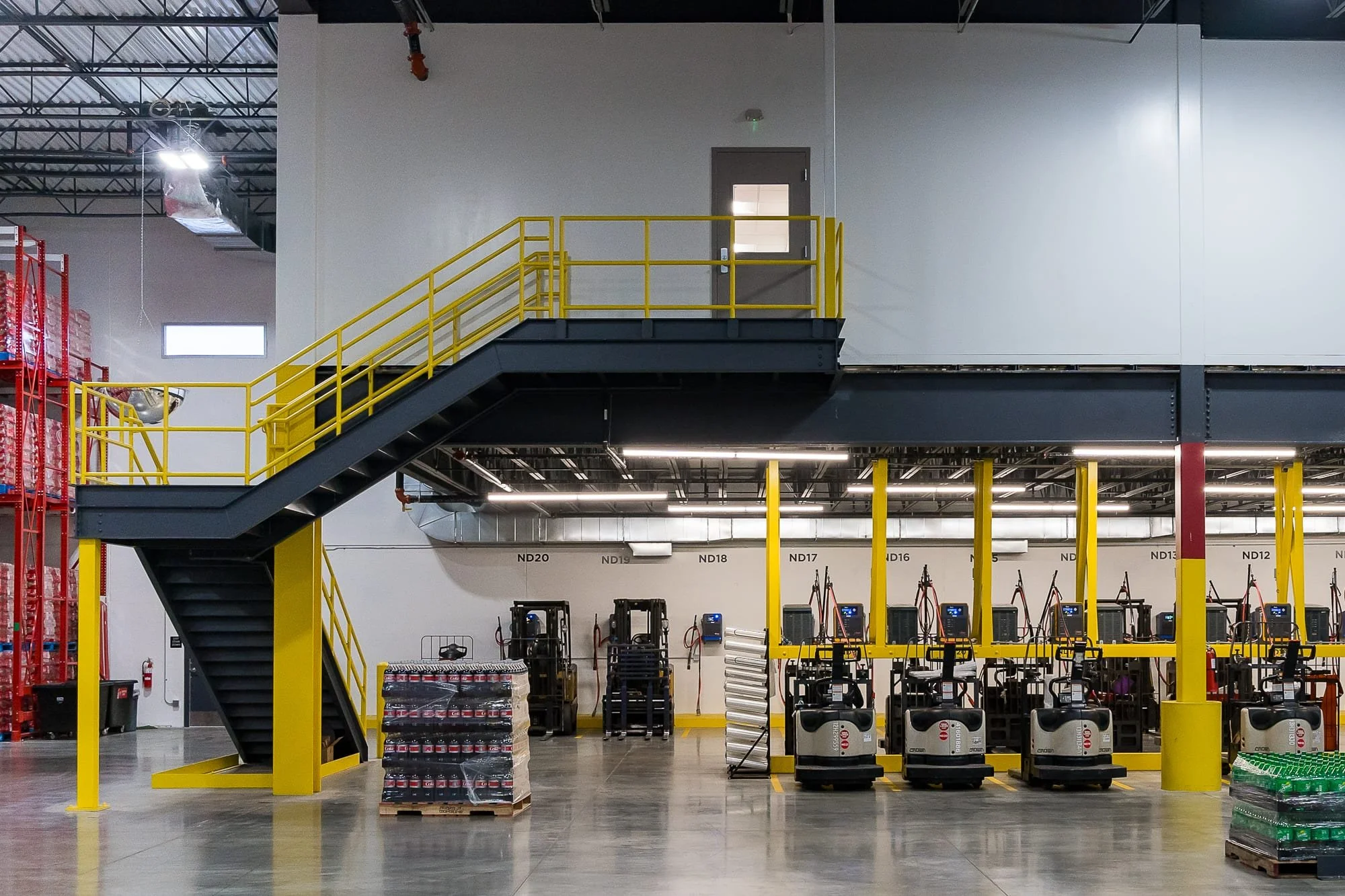 Interior of a clean industrial warehouse with yellow safety railings, a raised mezzanine, forklifts, palletized beverages, and polished concrete floors.