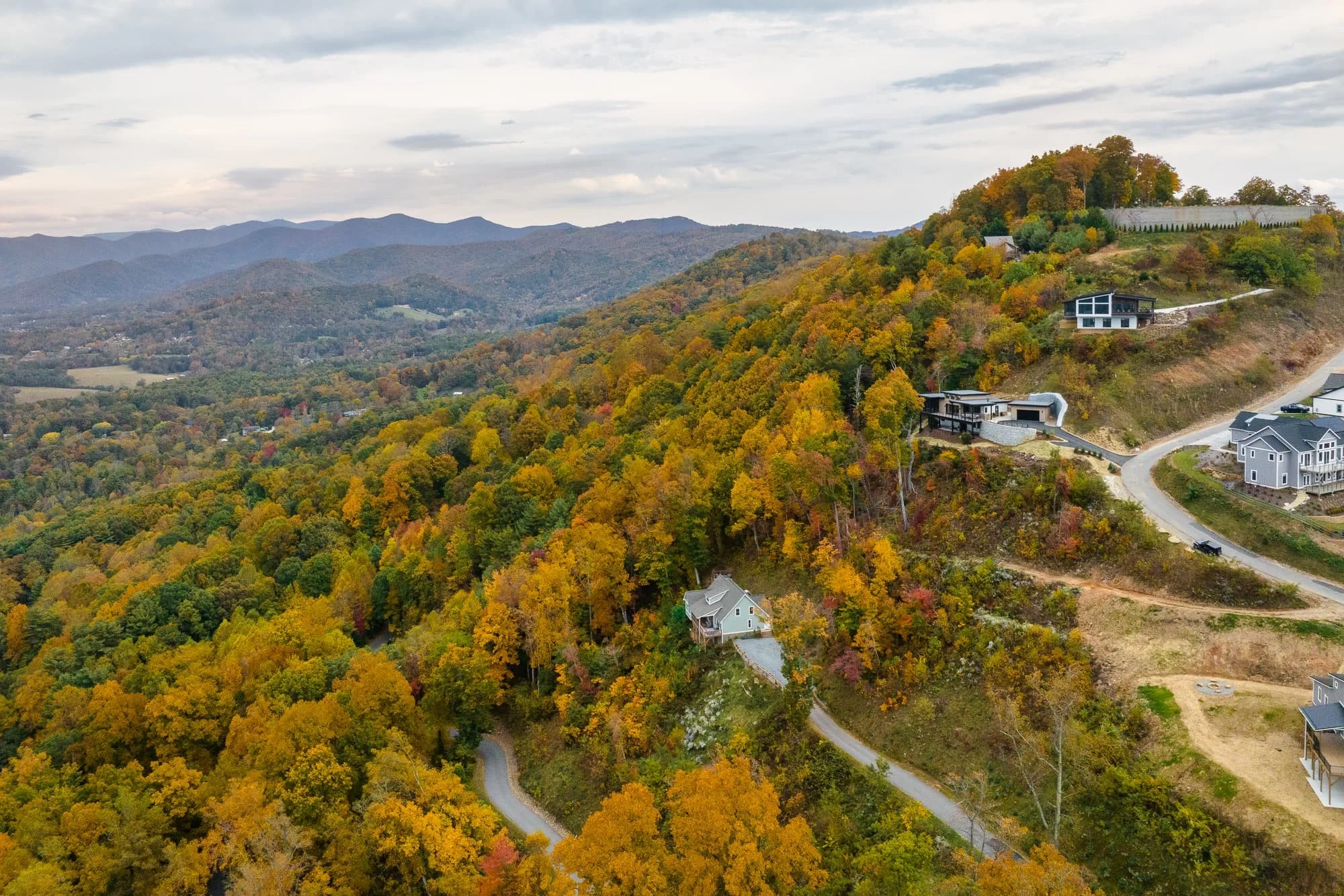 Aerial view of hillside homes among colorful fall trees, winding roads, and layered mountain views.