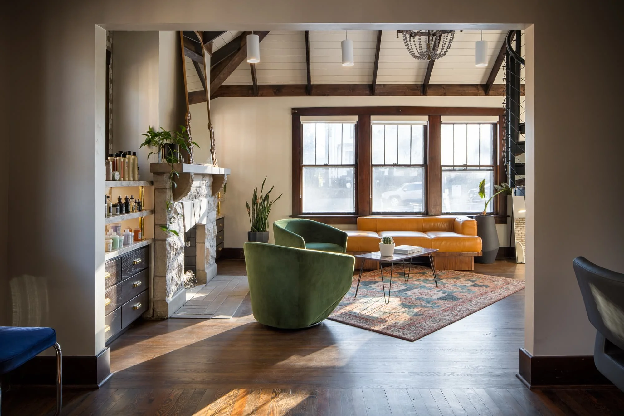 Framed view into a salon lounge with stone fireplace, green chairs, leather sofa, wood floors, and afternoon light.