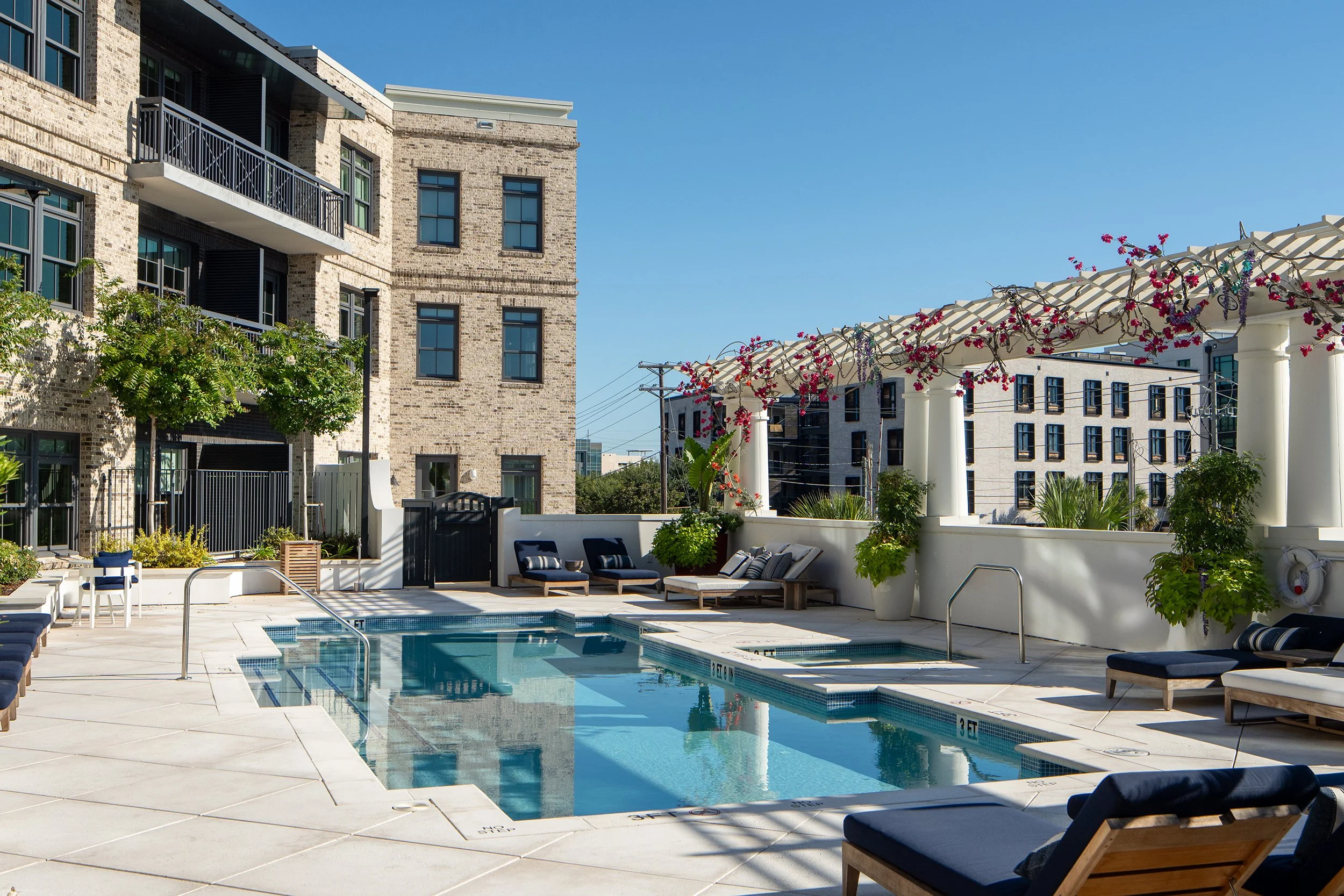 Outdoor pool deck with a primary lap pool and adjacent spa, a white pergola draped in flowering bougainvillea, navy and wood loungers, and brick building facades flanking the courtyard under a clear sky.