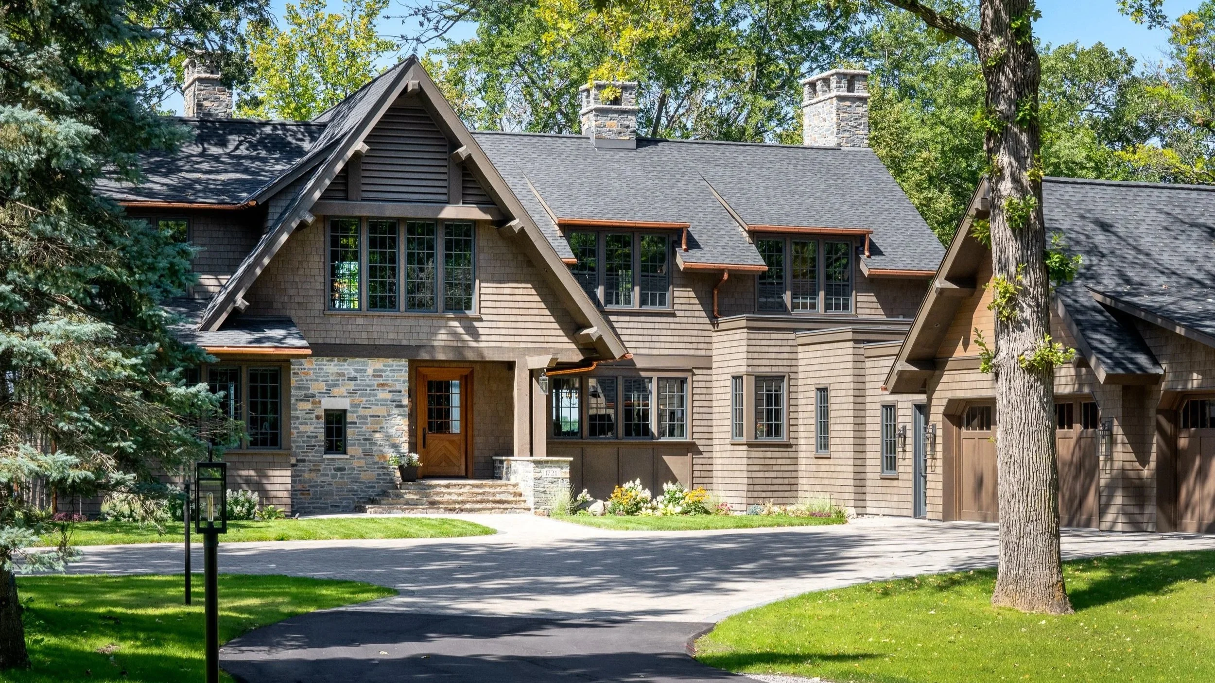 A large modern house with a combination of stone and wooden exterior, multiple windows, and a dark shingled roof, surrounded by trees and a green lawn.