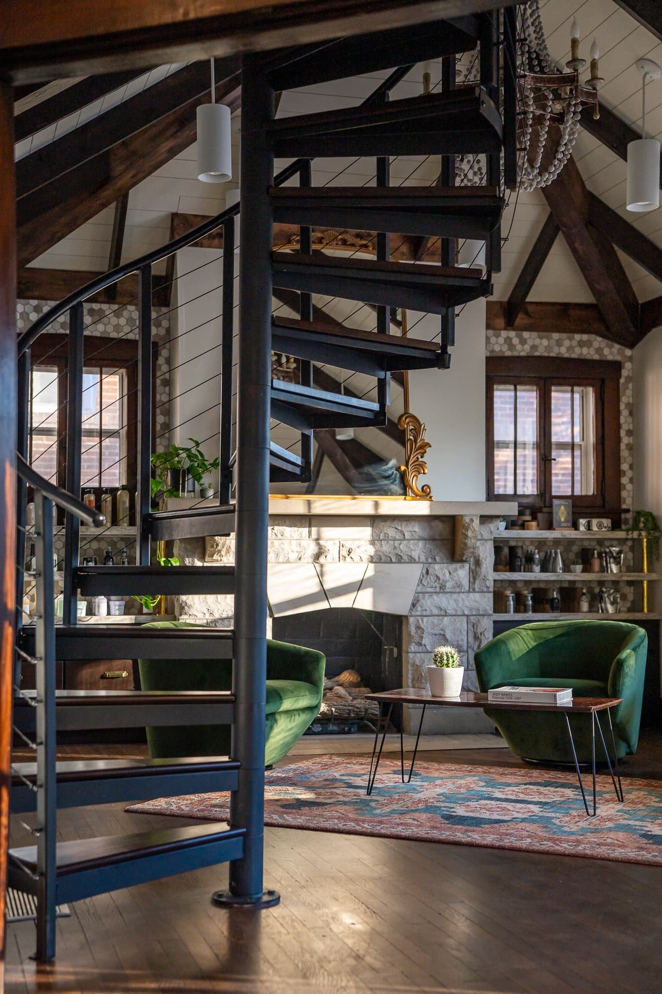 Black spiral staircase rises above a salon lounge with vaulted wood beams, stone fireplace, and green seating.