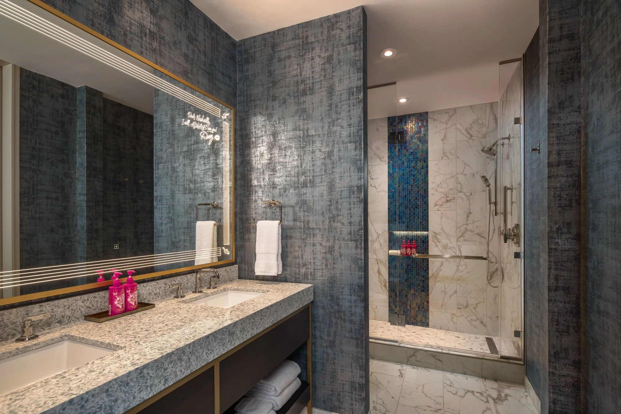 Blue hotel bathroom with a double vanity, illuminated mirror, marble walk-in shower, and mosaic tile details.