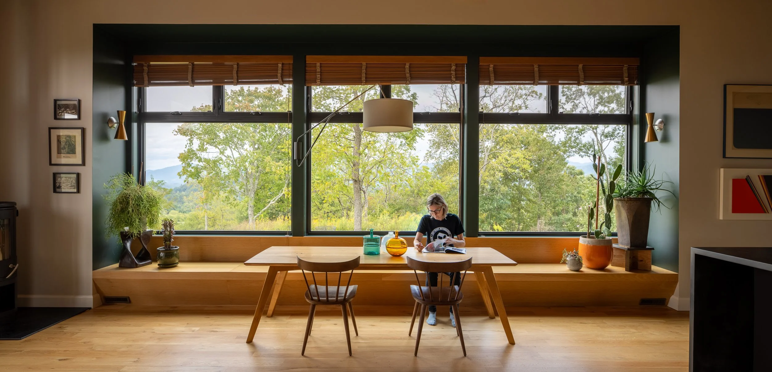 A woman sitting at a wooden dining table near large window with a view of trees and mountains, reading a magazine surrounded by decorative vases and plants in a modern living space.