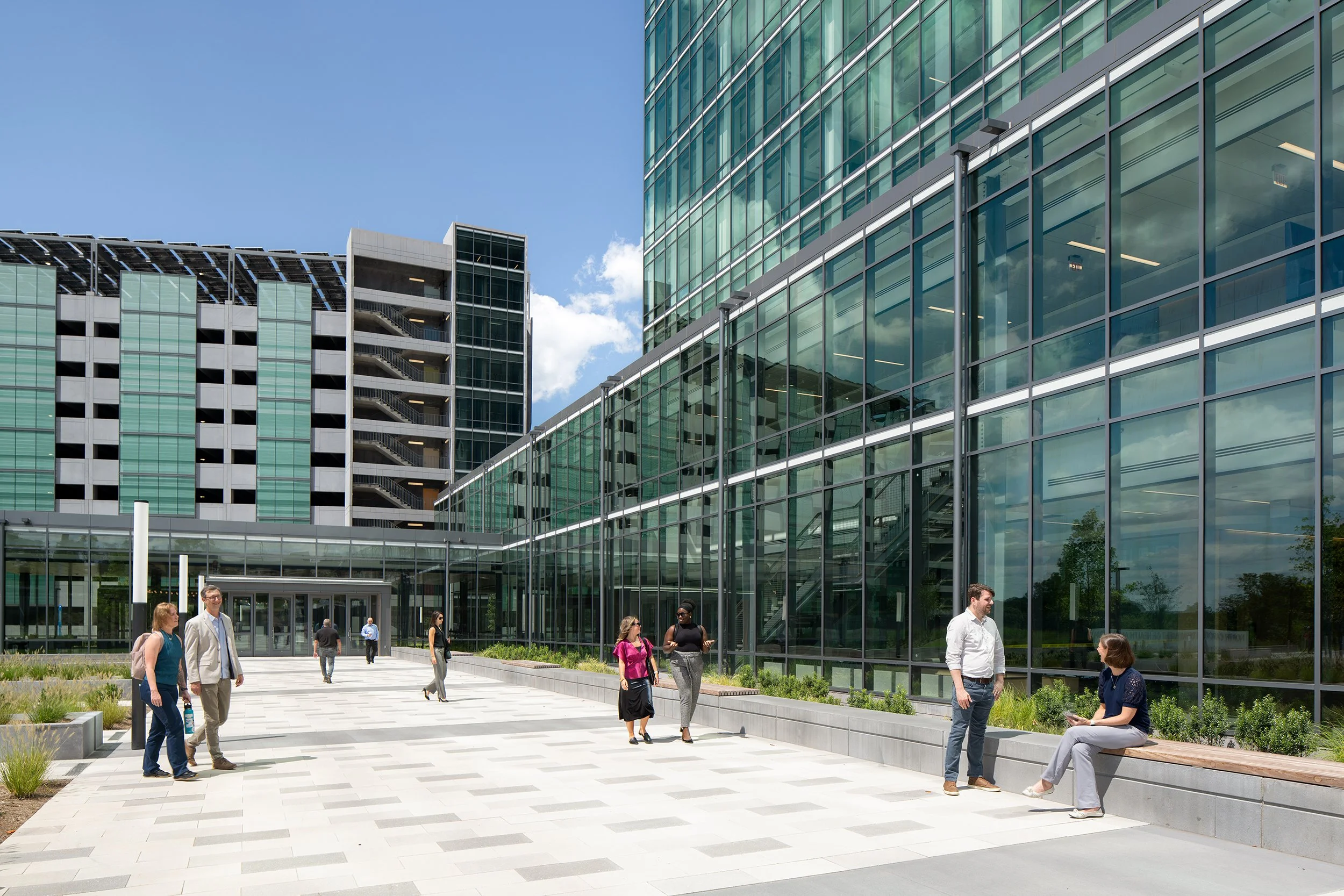 Plaza view between the office tower and the adjacent parking deck, with visitors walking past raised native plantings and integrated bench seating along the glass concourse.