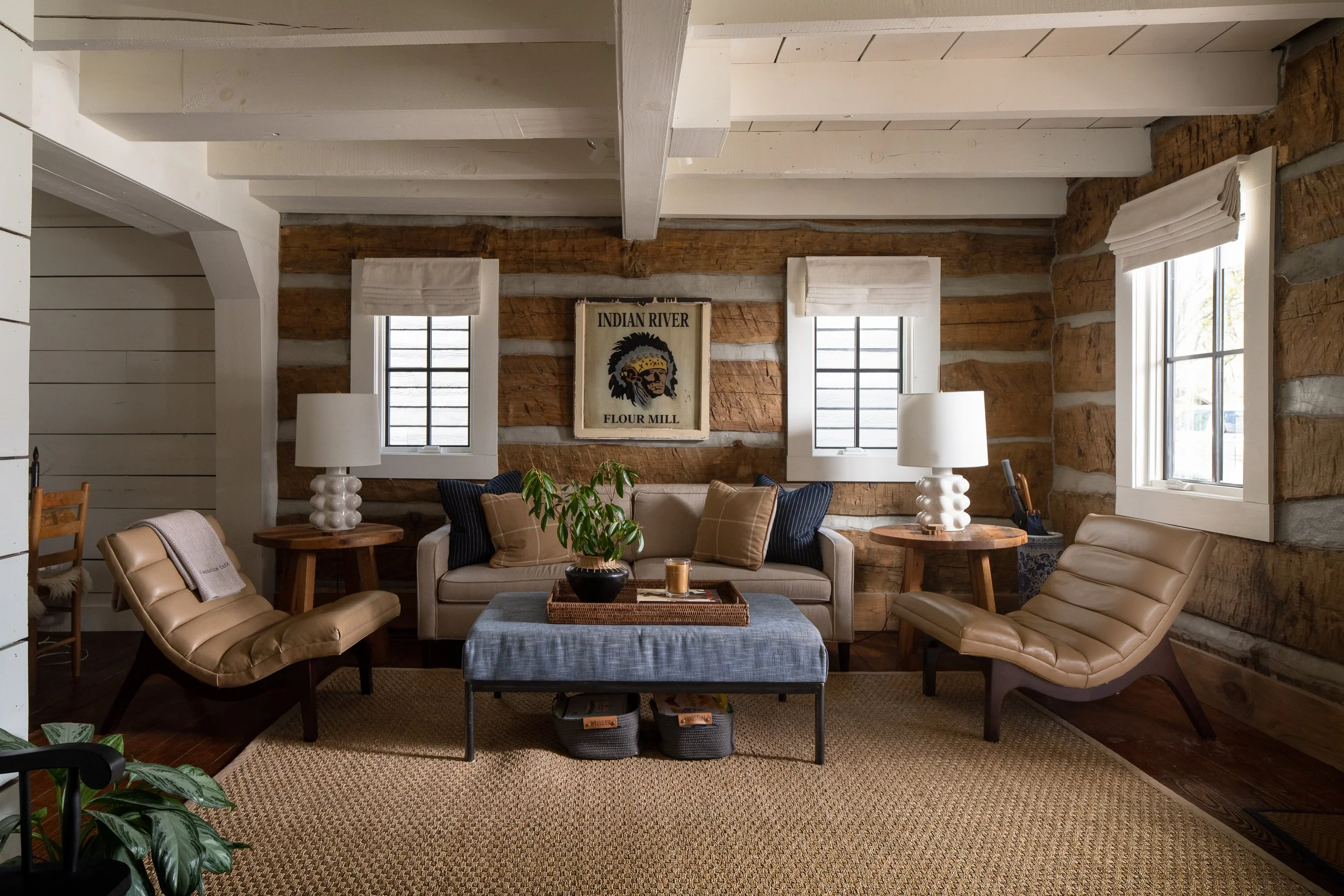 Rustic living room with log walls, white ceiling beams, layered seating, and neutral furnishings.