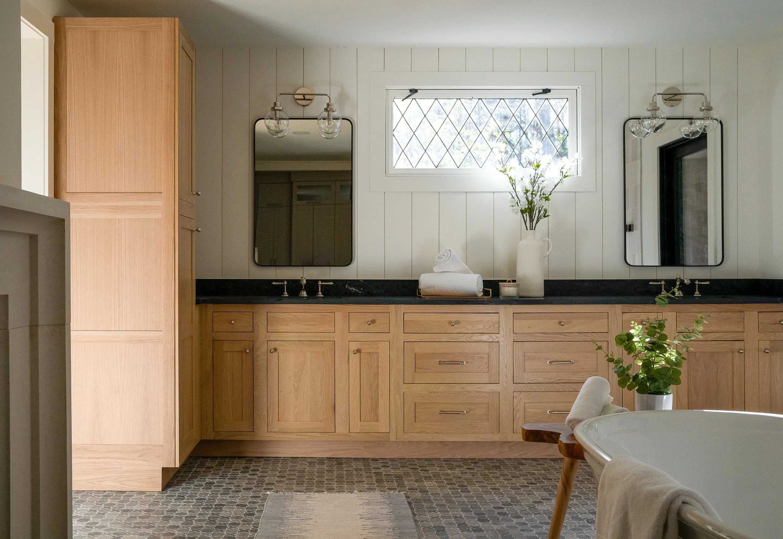 Primary bathroom with oak double vanity, black countertop, wall mirrors, globe sconces, and patterned floor tile.