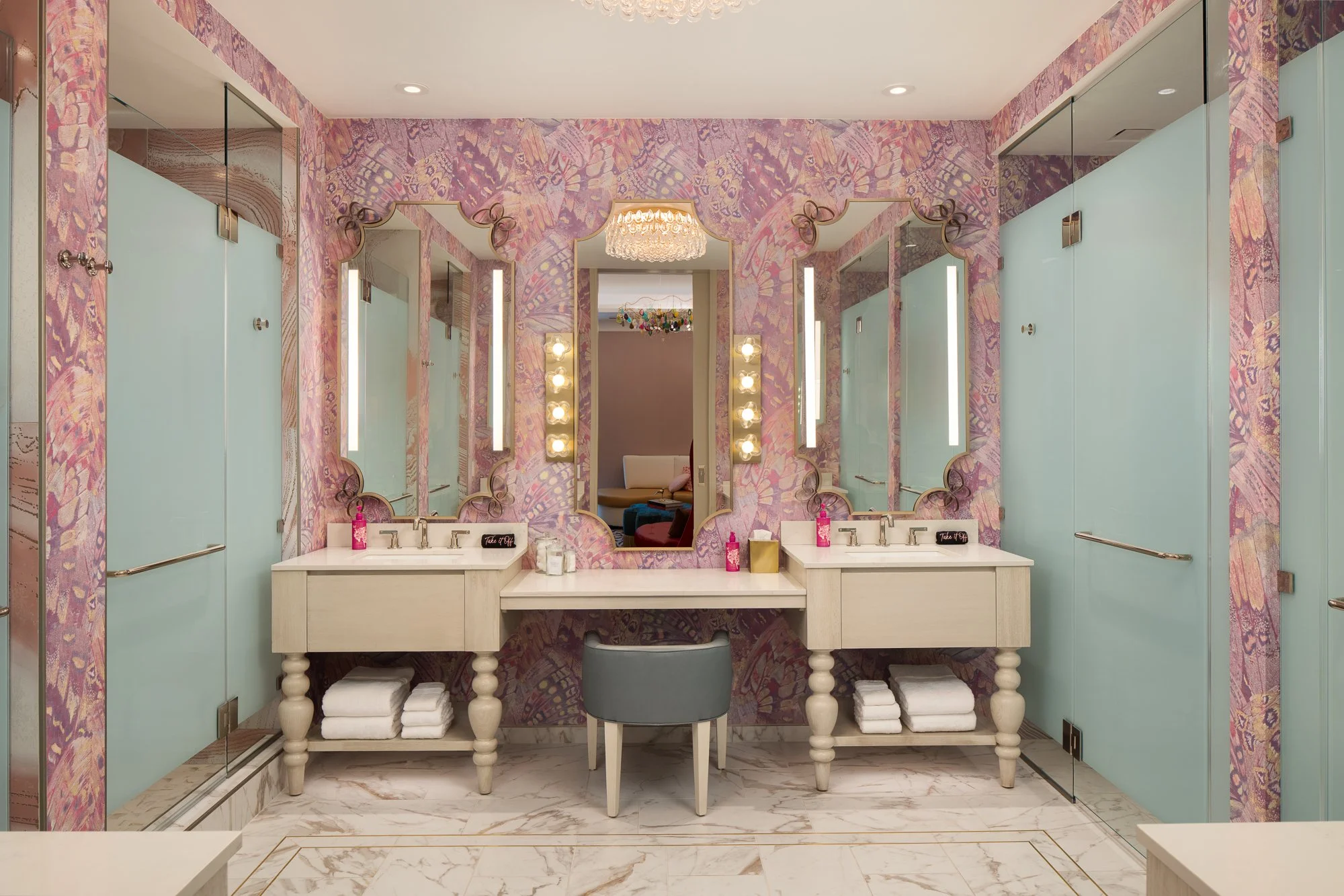 Symmetrical vanity room with patterned pink walls, decorative mirrors, dual sinks, frosted glass doors, and marble flooring.