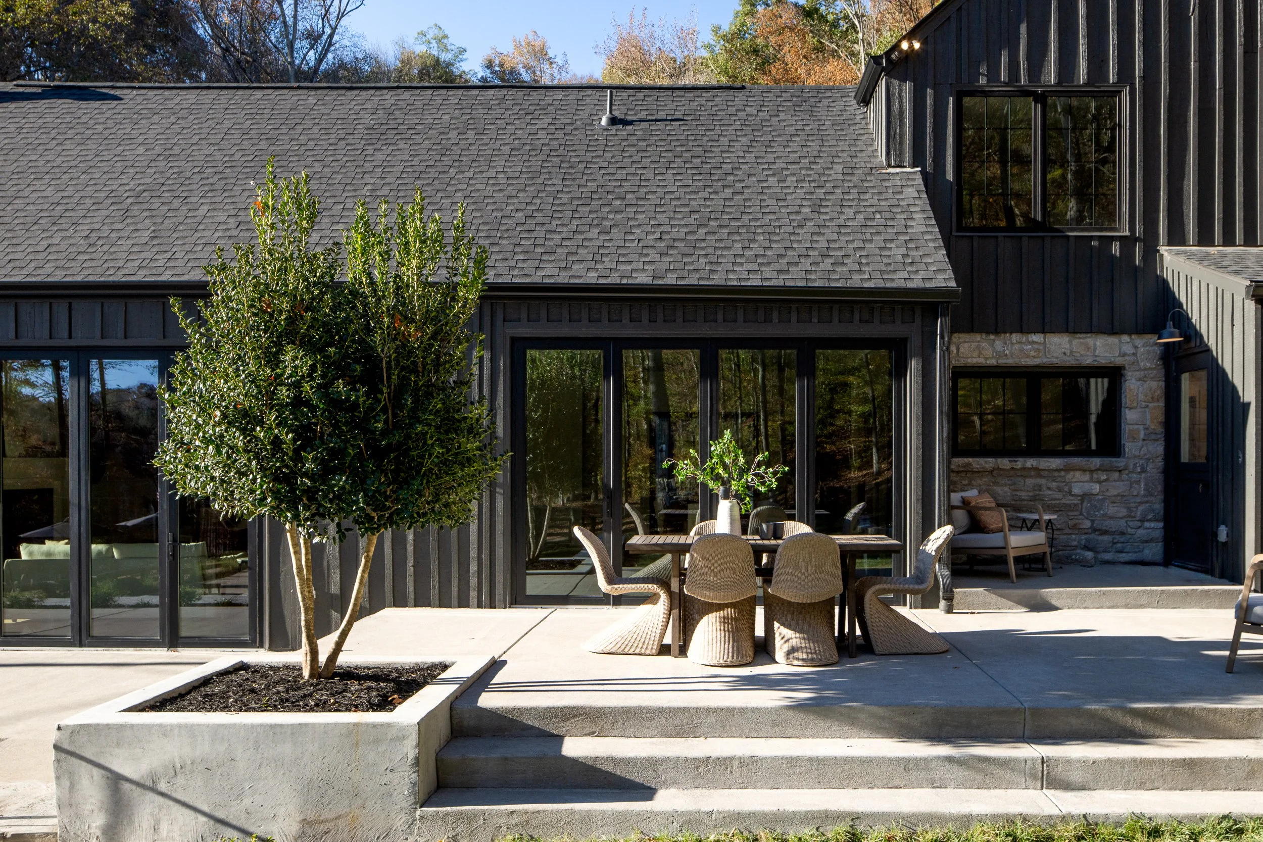 A modern house with black exterior walls, large windows, and a gray shingle roof. There is a small outdoor dining area with a round table and wicker chairs on a concrete patio. A young tree is planted in a rectangular bed in the foreground.