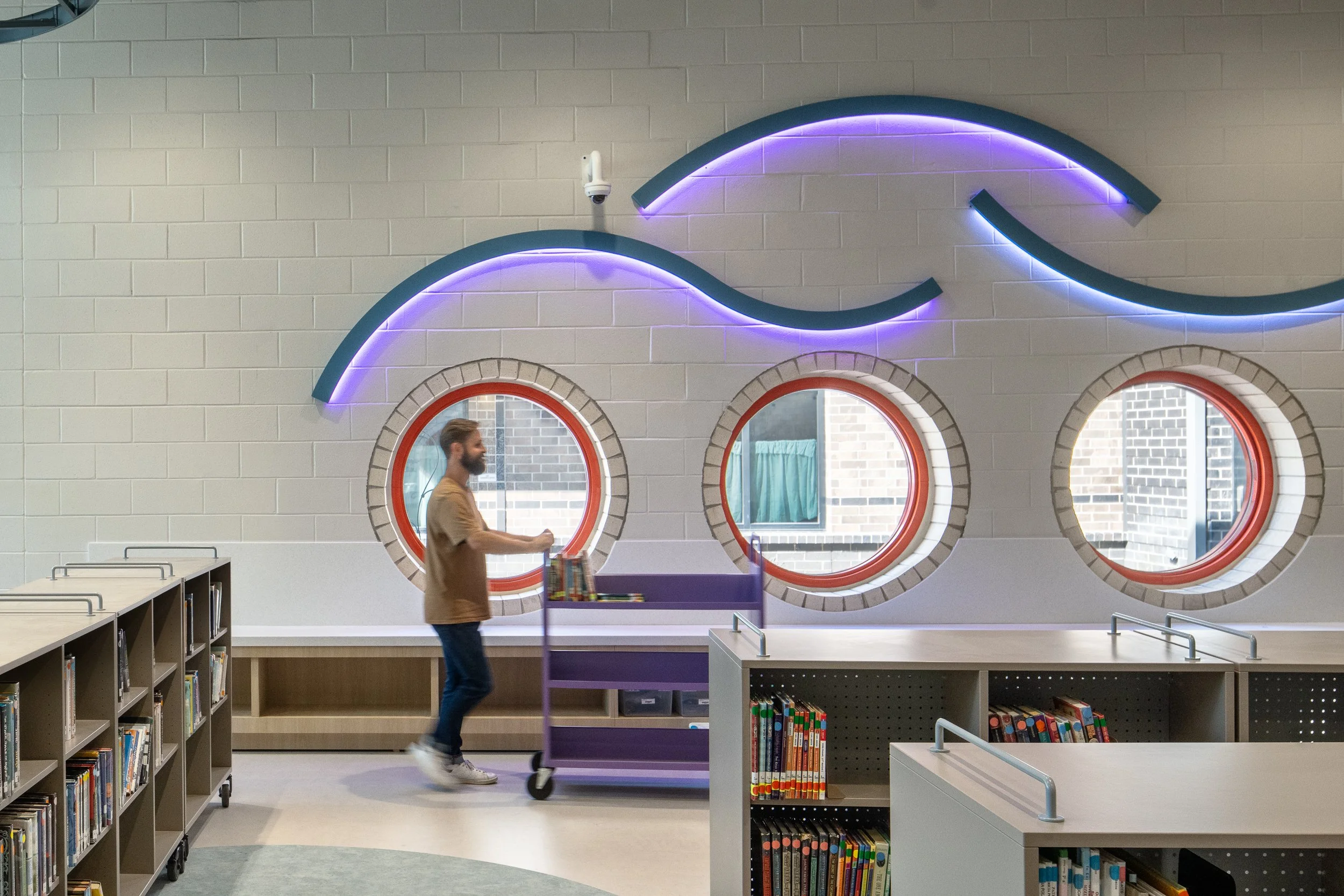 Library wall detail with glowing wave forms, round red-framed windows, bookshelves, and a person pushing a book cart.