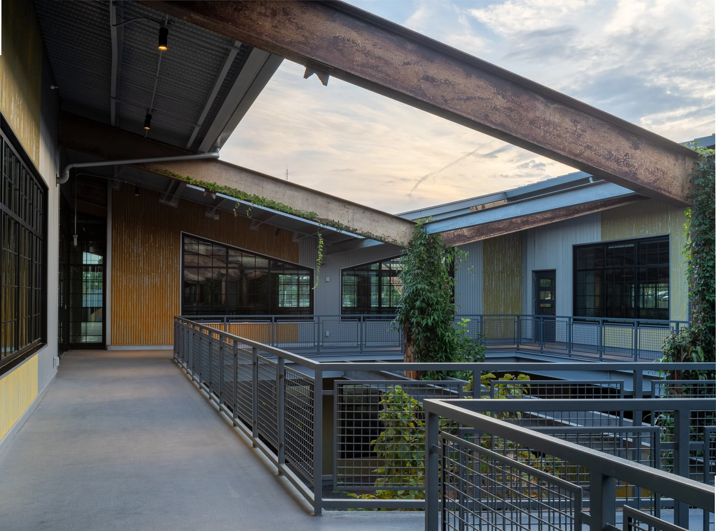 Open second-floor walkway beneath an original rusted steel beam, with yellow ghost-painted corrugated metal walls, climbing vines on the structural columns, and views back to the courtyard at dusk.