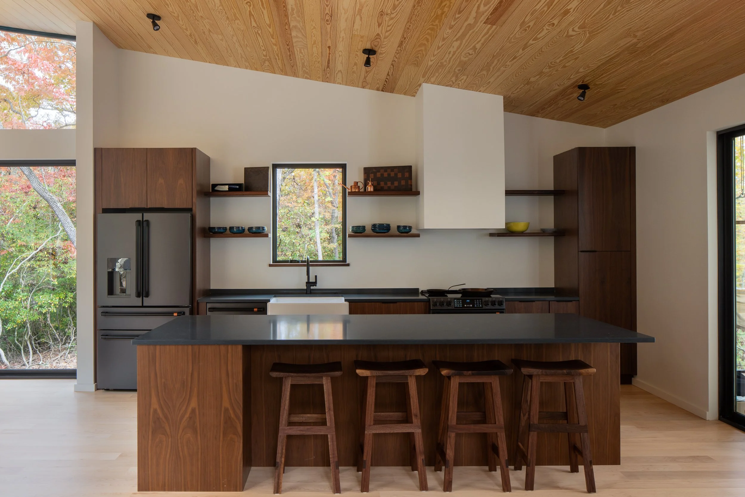 Kitchen with walnut cabinetry, dark countertops, wood stools, and a warm wood ceiling.