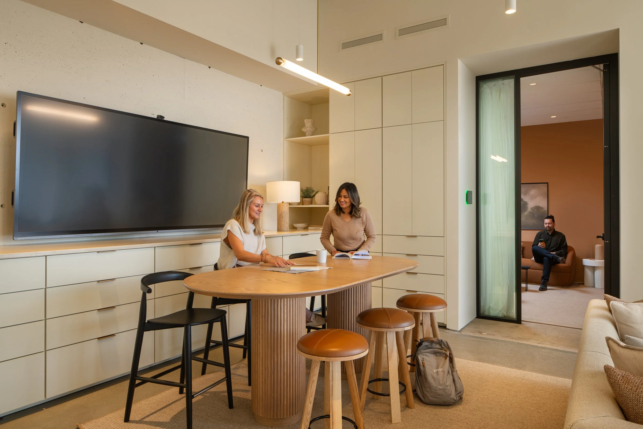 Two women sitting at a wooden conference table, smiling and talking. There is a man sitting in a room background on a couch, looking at his phone. The room has a modern decor with a large black TV, a white cabinet, a lamp, and a sliding glass door.