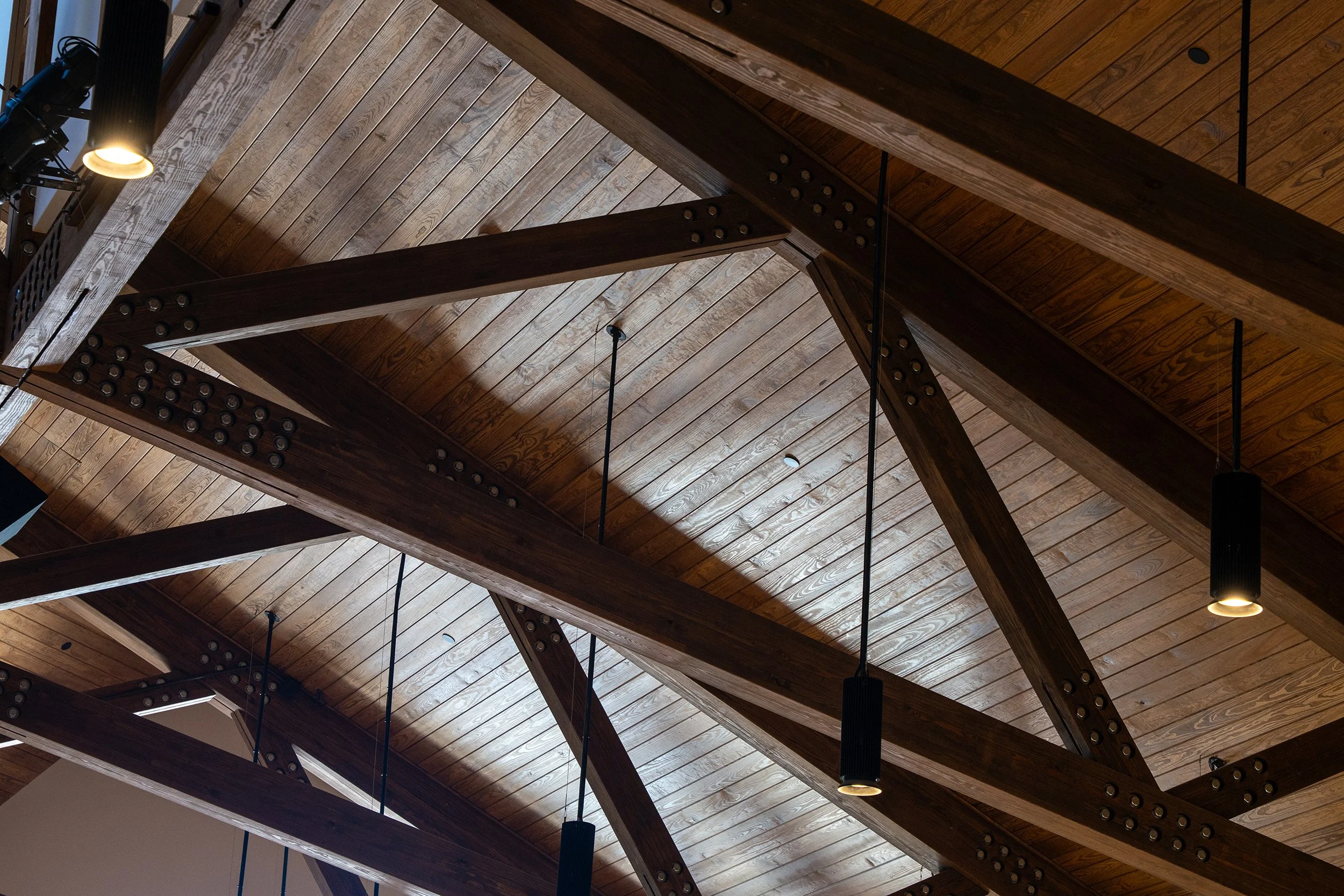 Detail of exposed heavy timber roof trusses with bolted steel connections and pendant lights at Storyhill Church in Davidson, NC.