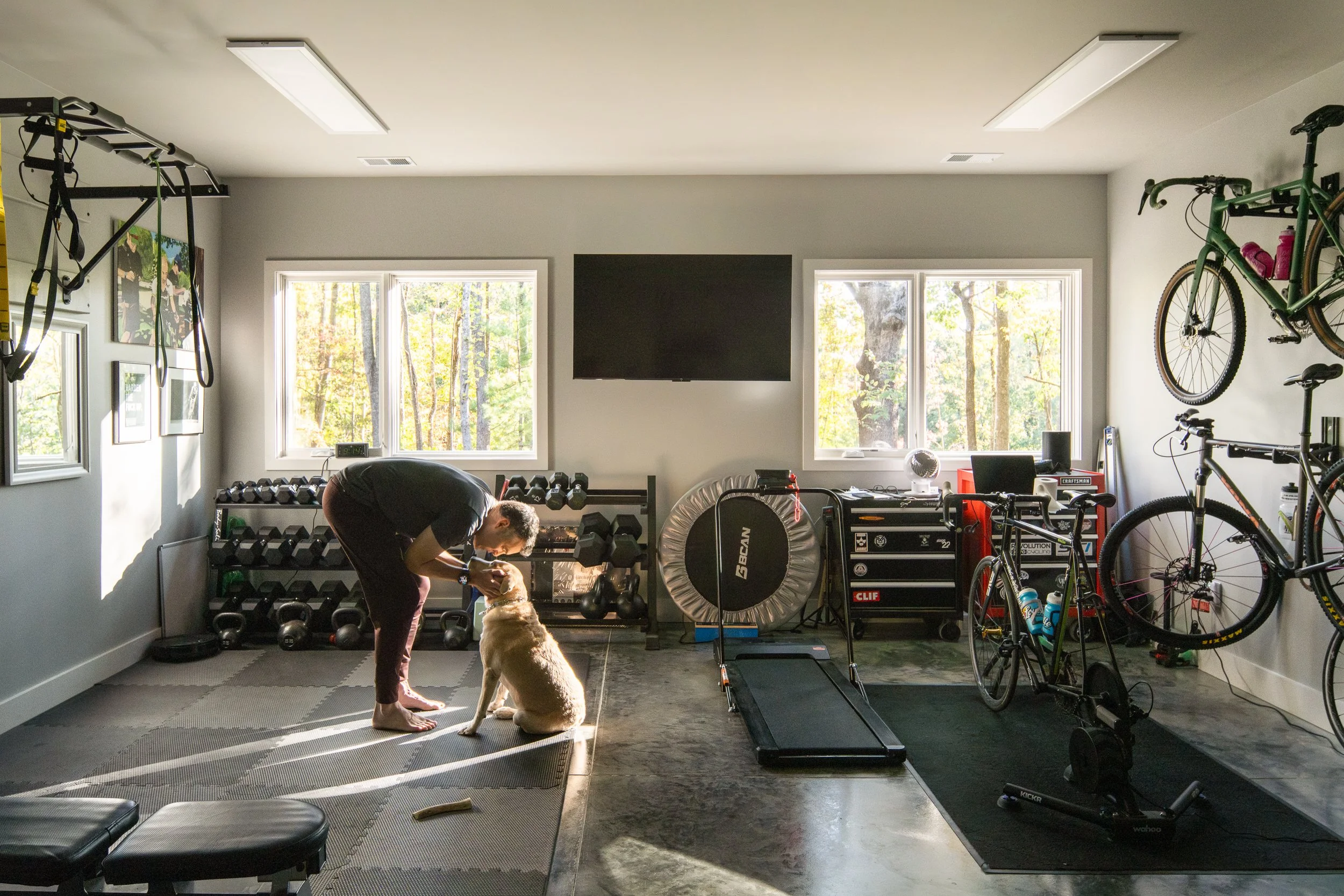 Home gym with weights, bikes, and a client bending down to greet Willet.
