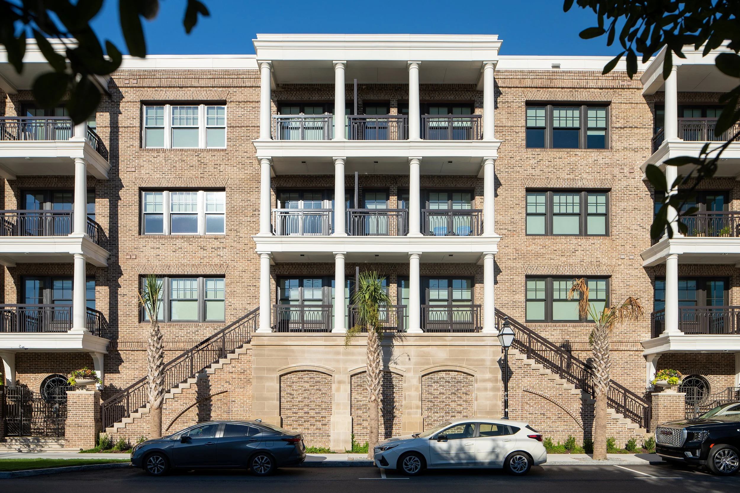 Symmetrical facade of the brick residential building with a raised central entry flanked by curving paired stairs, full-height white columns spanning three stories, wrought iron balcony railings, and palmetto palms in the courtyard.