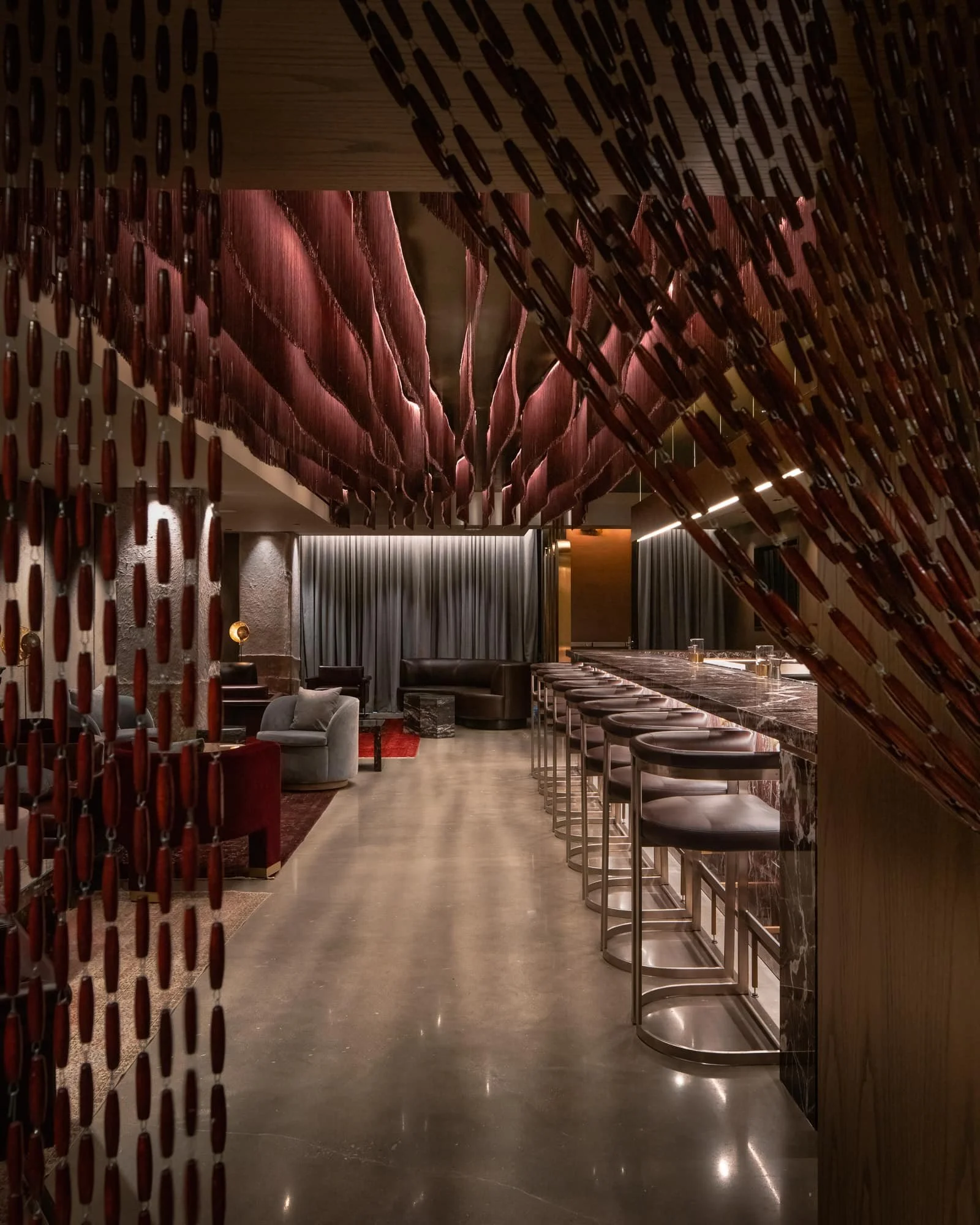 View into a luxury bar interior framed by decorative beadwork, marble bar seating, polished concrete floors, and burgundy ceiling forms.