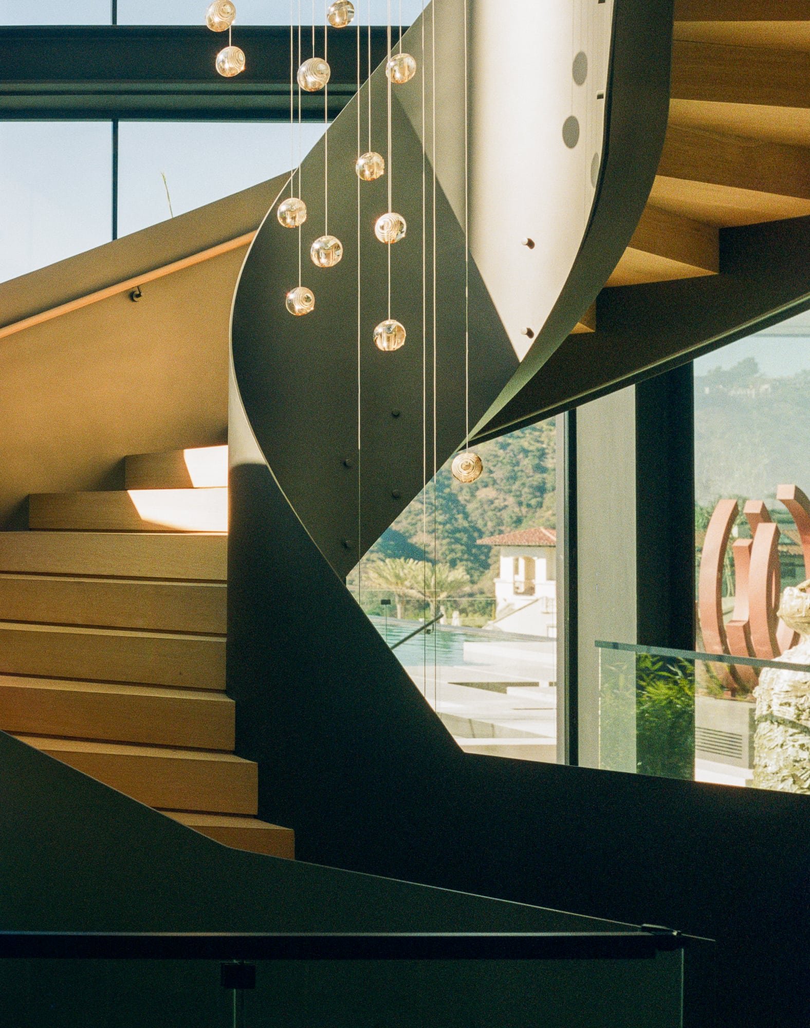 Sculptural stair with curved dark railing, pendant lights, wood treads, and hillside views beyond the glass.