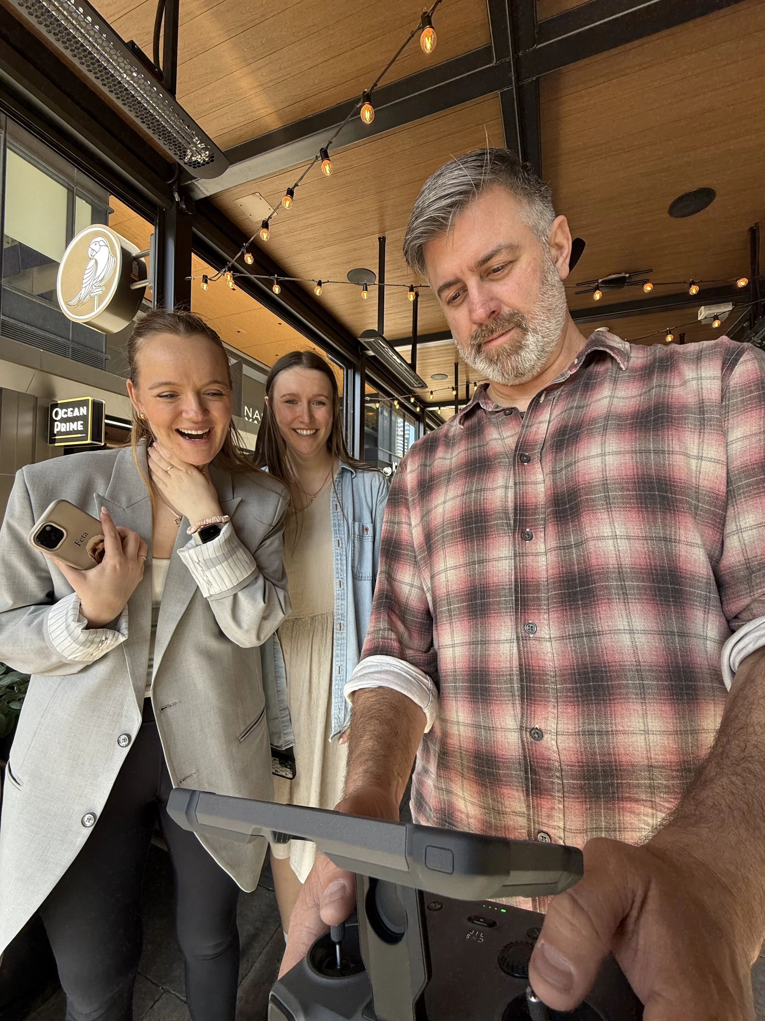 Three people looking at a drone remote controller, inside a cafe with wooden ceiling, string lights, and a digital menu board.