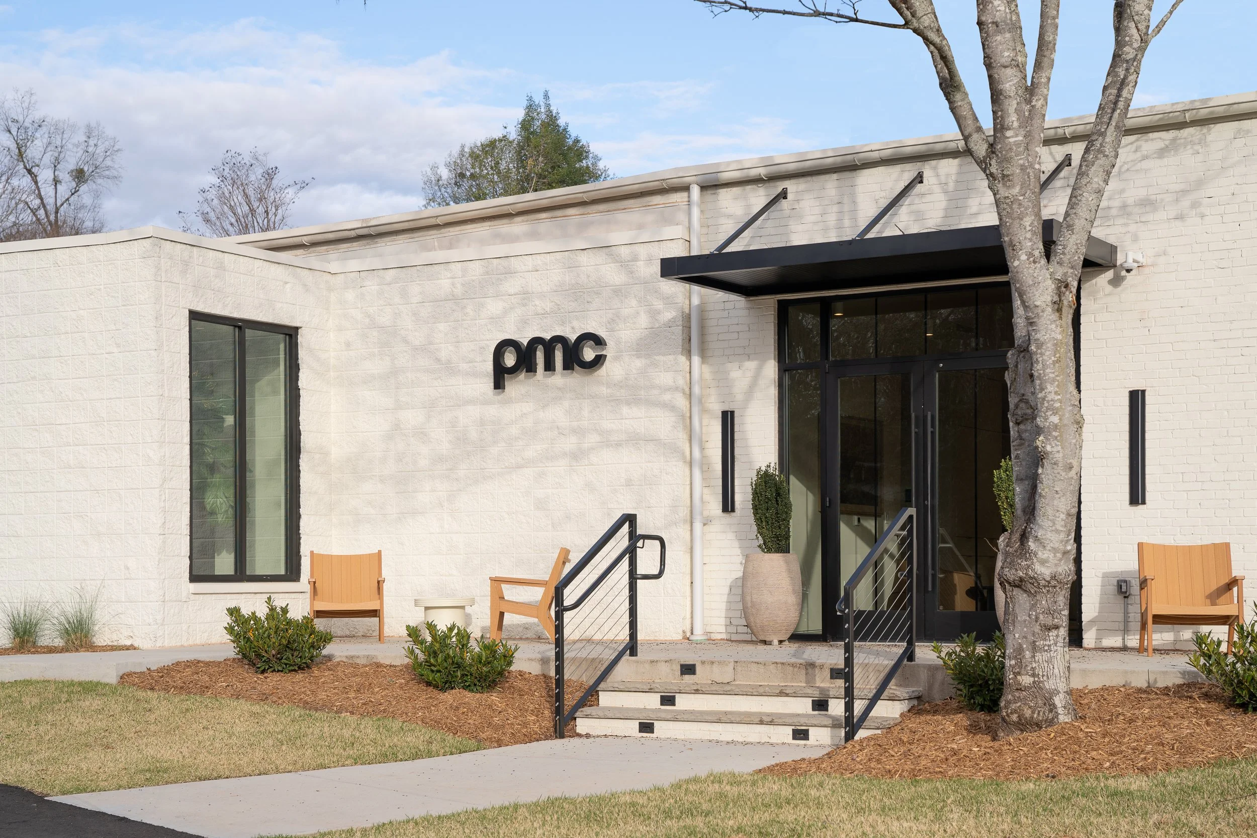 Front entrance of the PMC office with the lowercase brand signage mounted on white-painted brick, a cantilevered steel awning, cable-rail entry steps, and outdoor wood lounge chairs flanking the door.