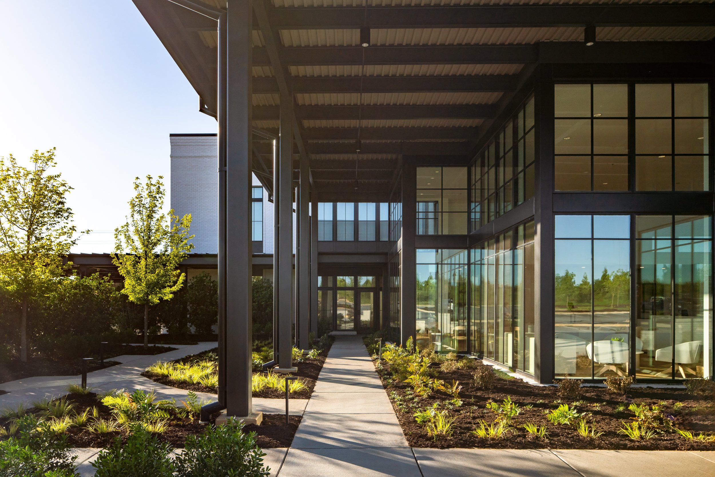 Modern building with large glass windows, surrounded by landscaped greenery and trees, with a pathway leading to the entrance.