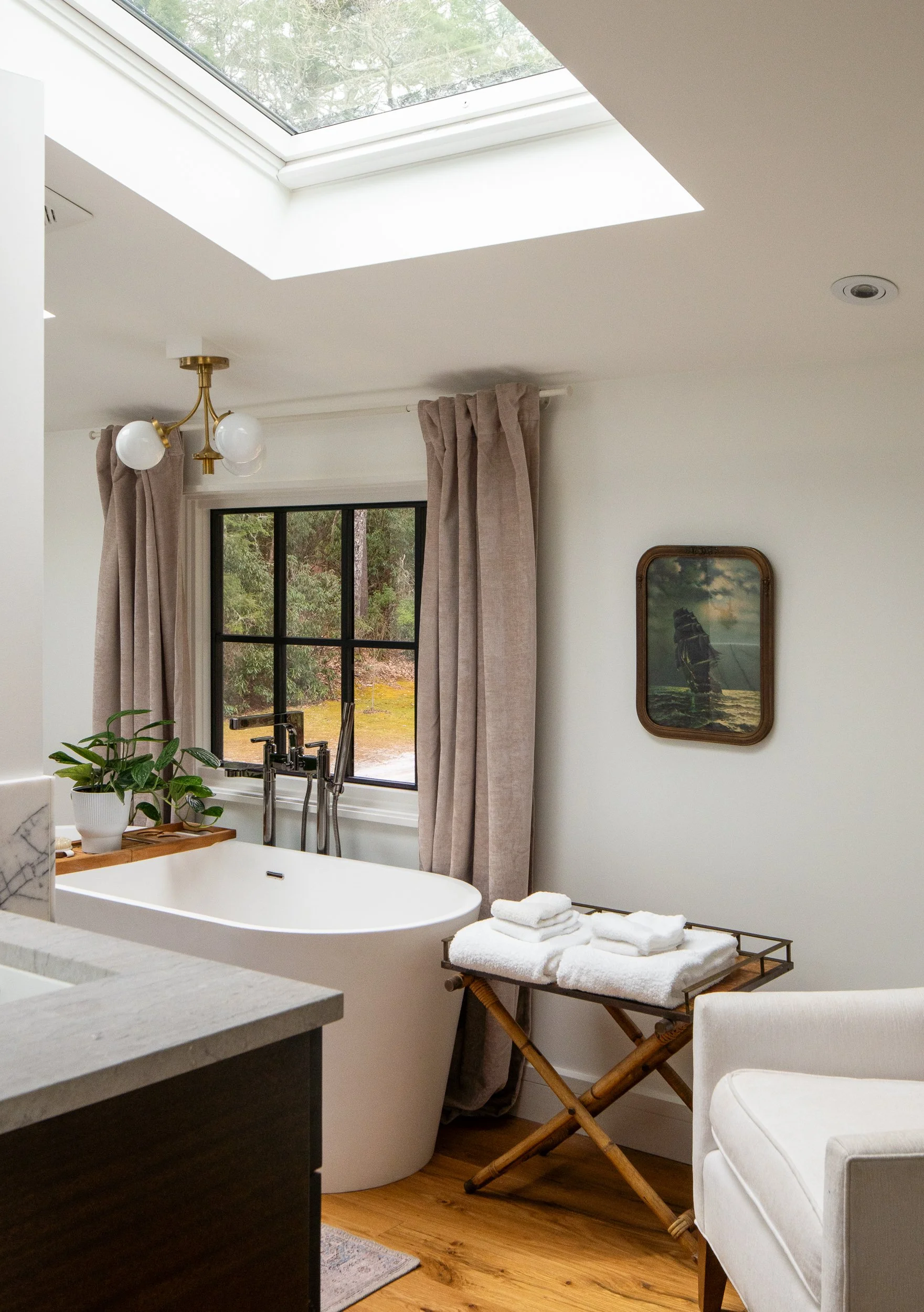 Bright bathroom corner with freestanding tub, skylight, linen curtains, folded towels, and mountain views.