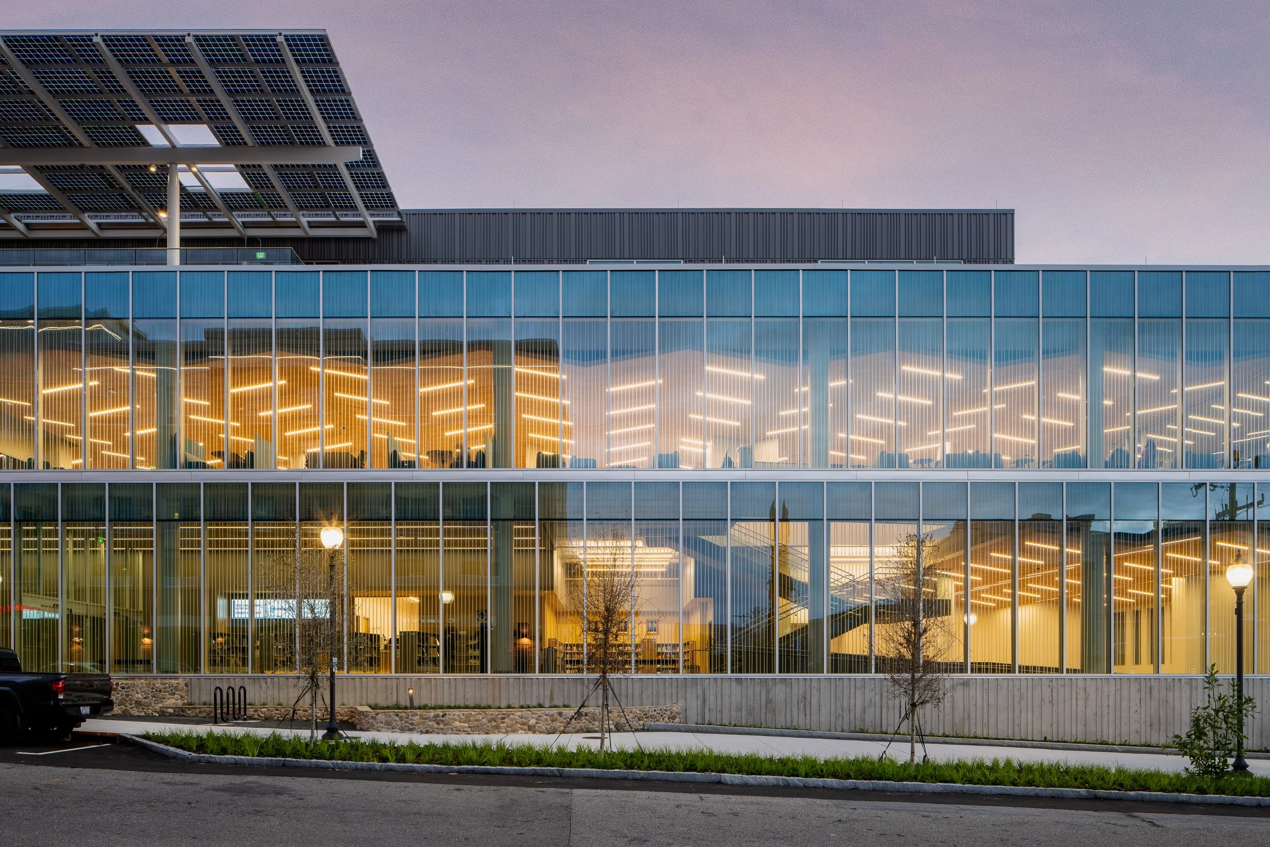 Glass-front library exterior at dusk with solar canopy, warm interior lighting, and modern facade.