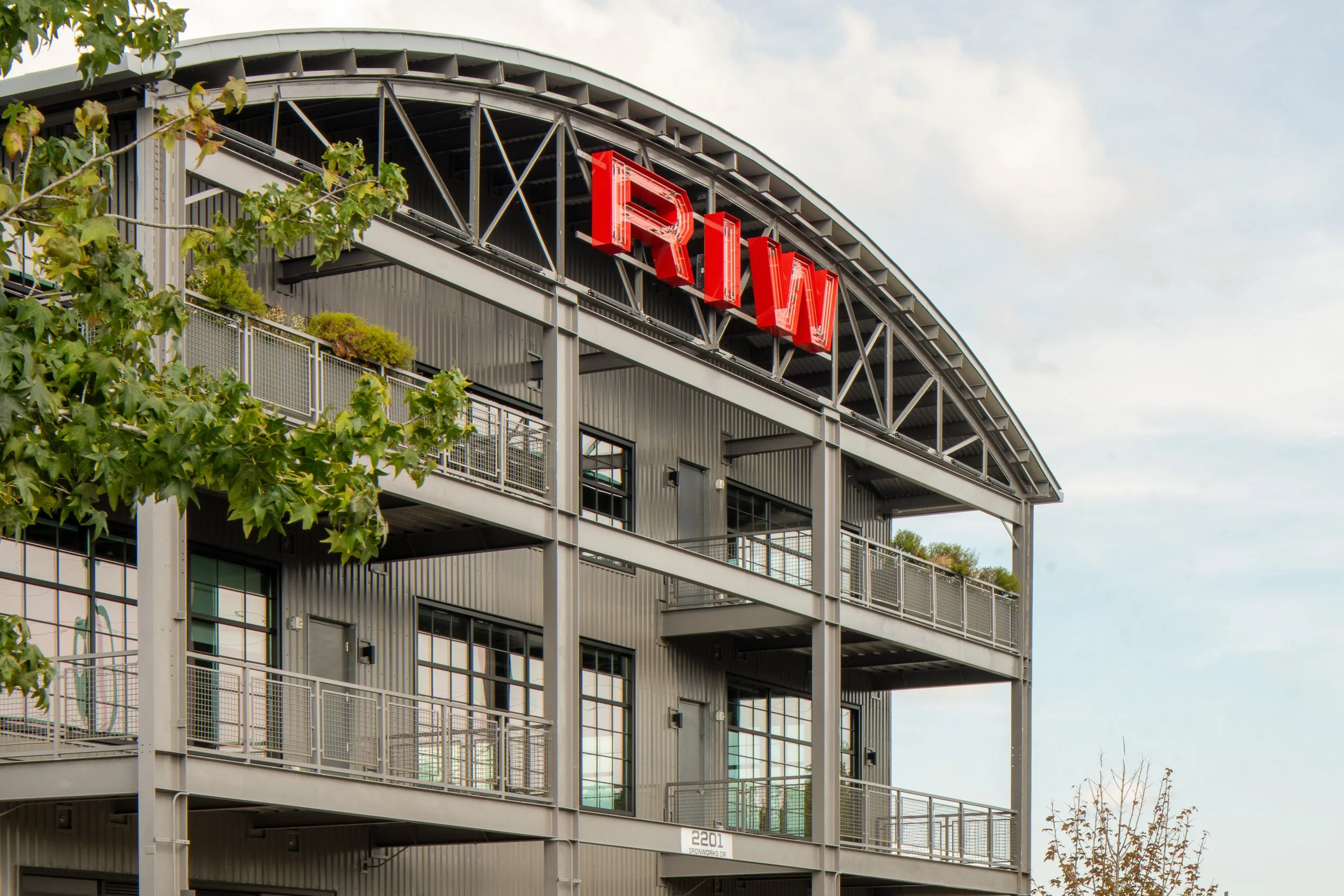Curved corrugated metal facade of the Raleigh Iron Works building with backlit red RIW lettering on the roof line, mesh-railed balconies, and tree branches framing the foreground.