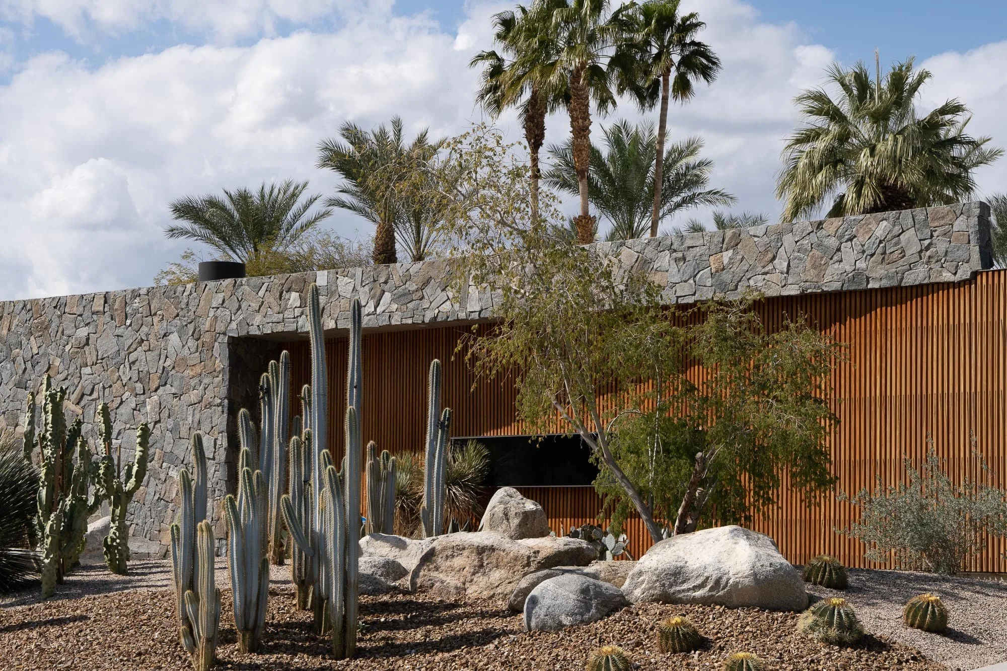 Desert residence with stone facade, vertical wood slat walls, cactus garden, boulders, and palm trees under a blue sky.