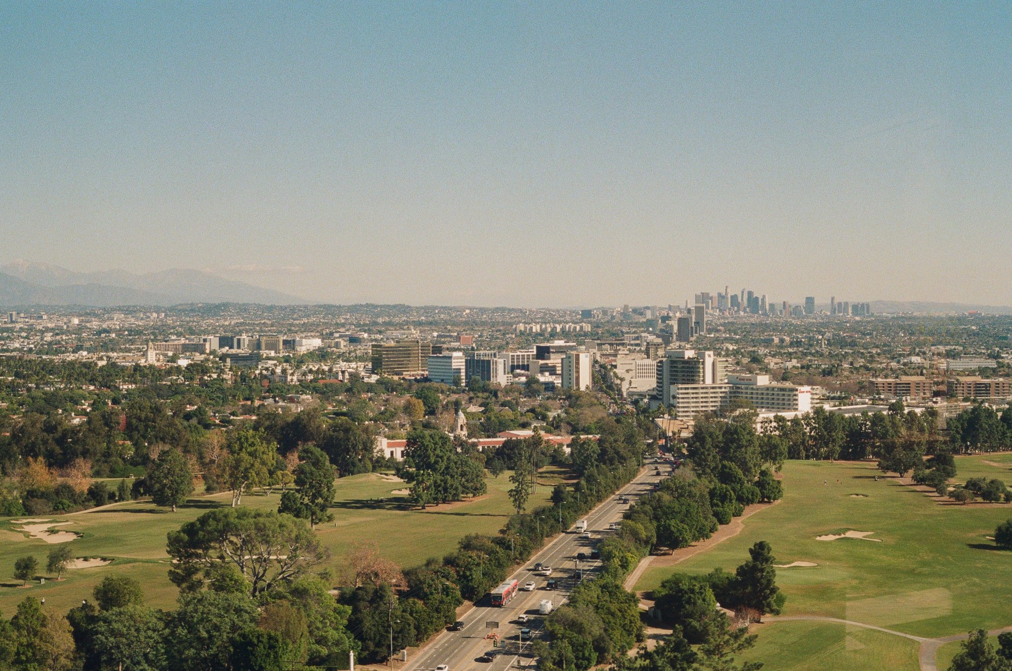 Elevated view across Los Angeles with a golf course, urban neighborhoods, and the downtown skyline in the distance.
