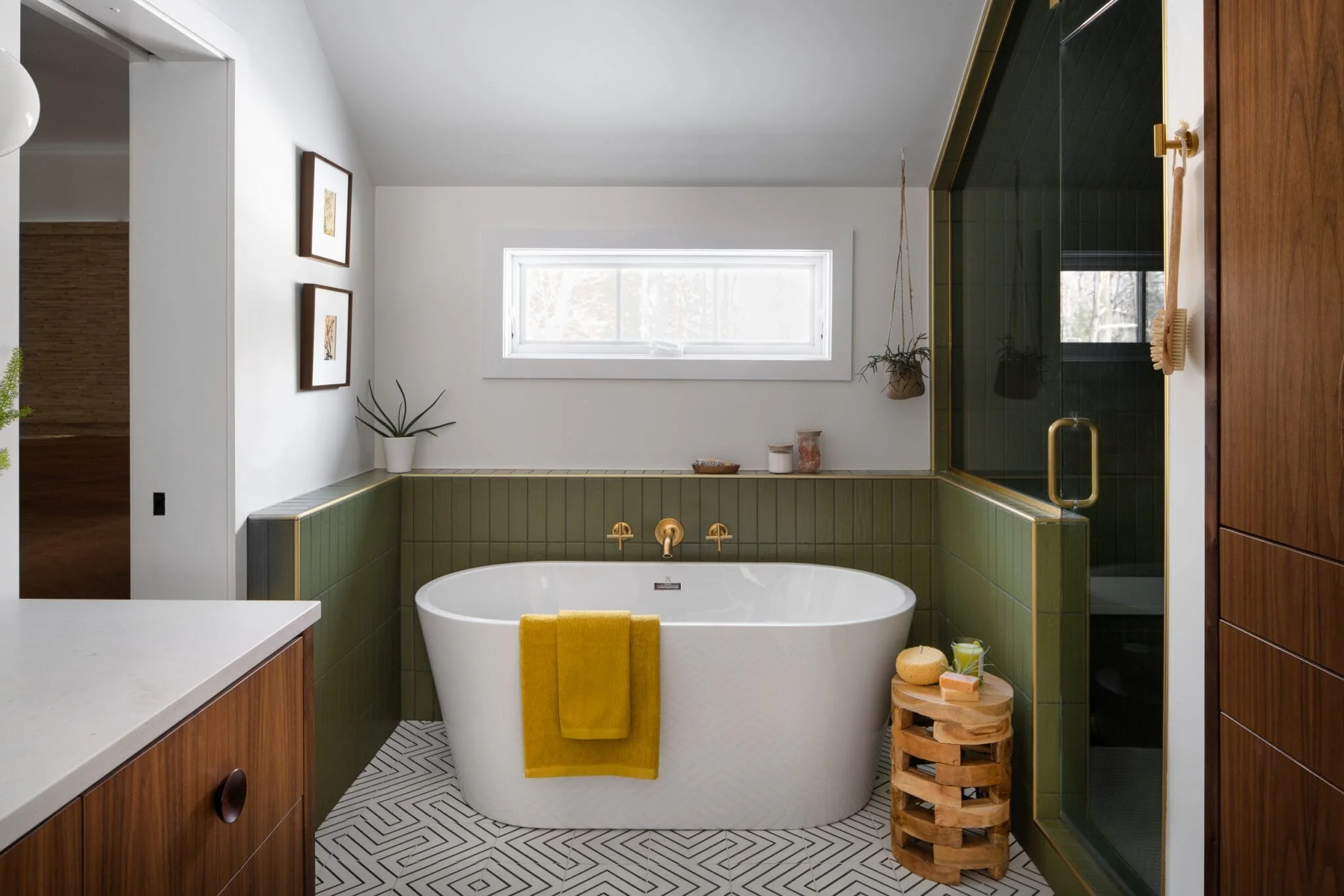 Primary bathroom with a freestanding soaking tub, olive green tile surround, brass wall-mount fixtures, geometric floor tile, walnut vanity, and framed artwork on the wall
