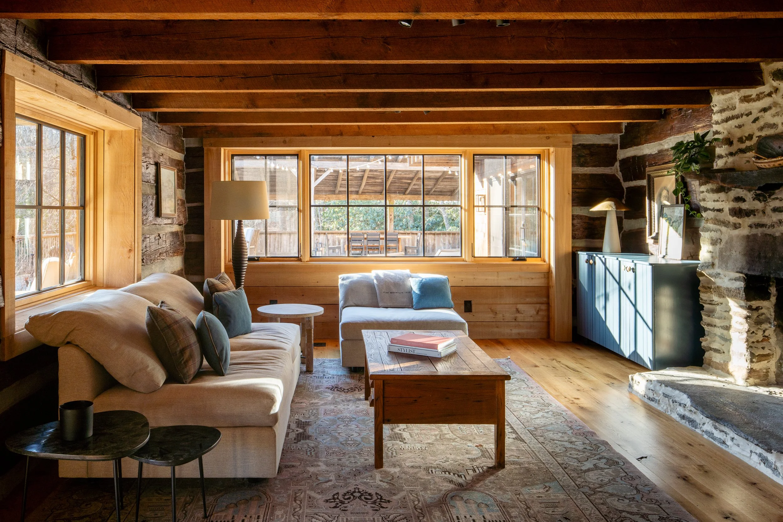 Cozy log cabin living room with sofas, wood beams, large windows, and a stone fireplace.