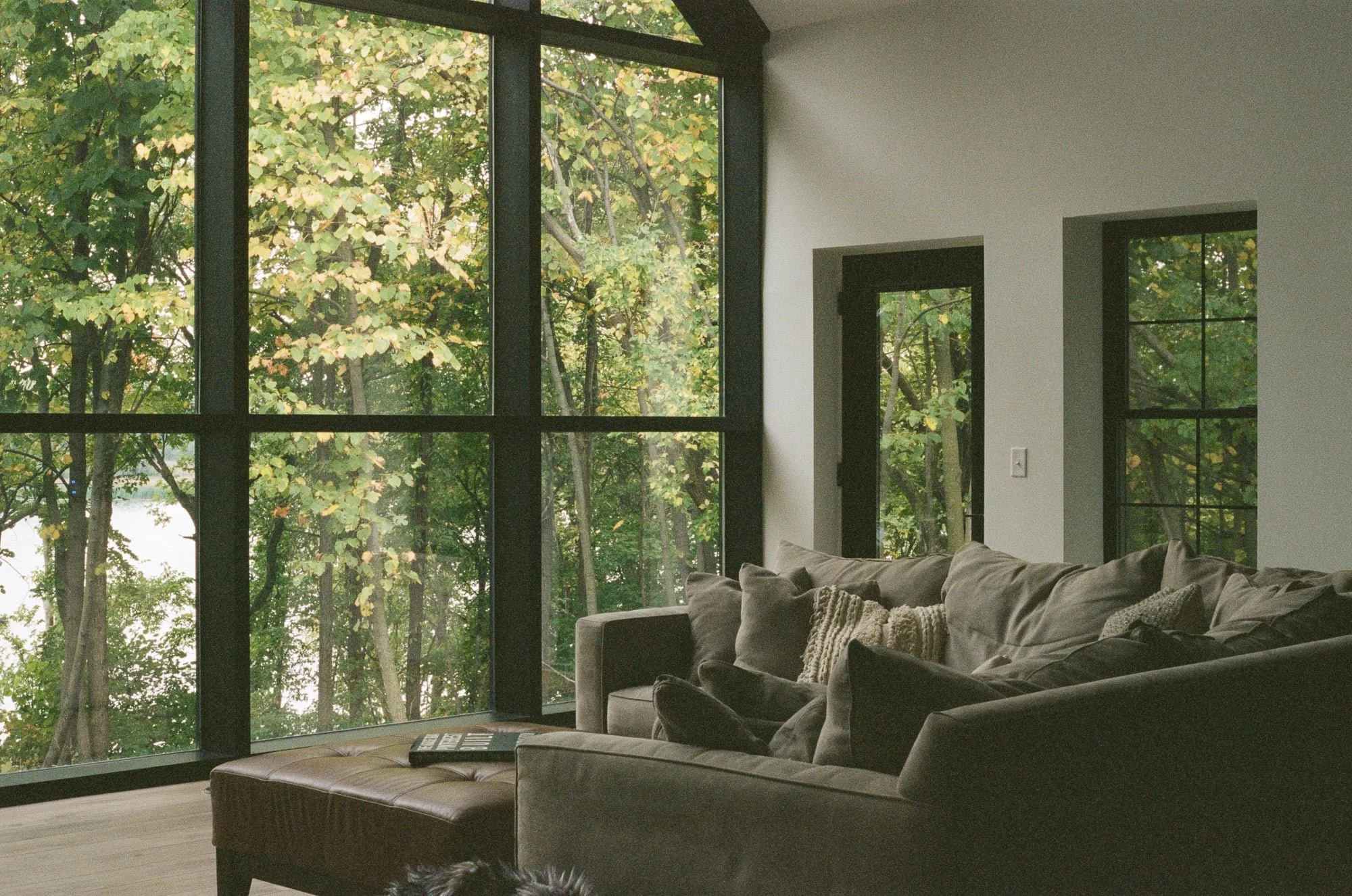 Living room with large windows showing green trees outside, tan sofa with pillows, and a remote control on a brown ottoman.
